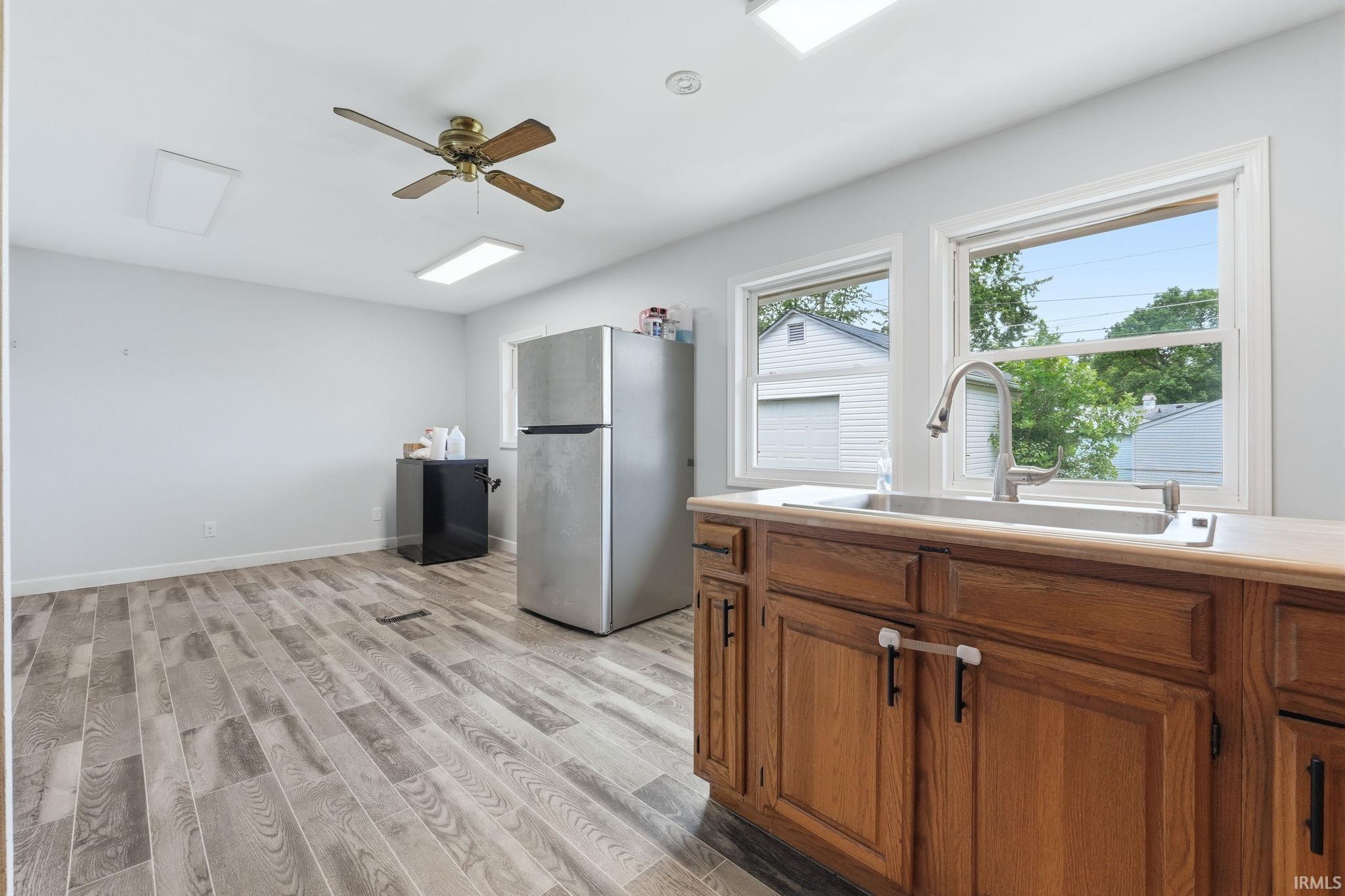 Kitchen area featuring wood-finish flooring, two large windows, a stainless steel sink with a high-arch faucet, wood cabinetry, and a ceiling fan