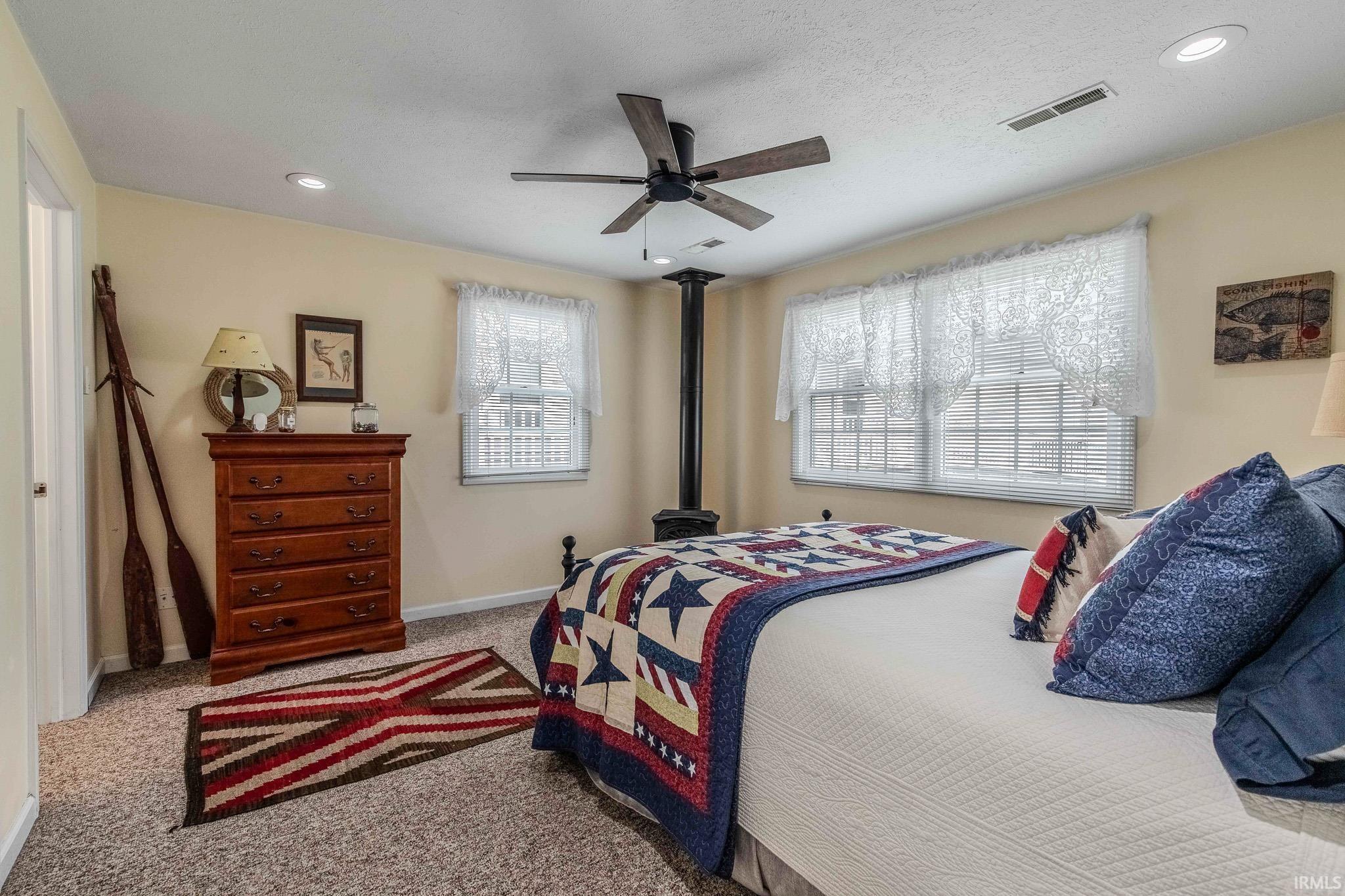 Carpeted bedroom featuring a ceiling fan, recessed lighting, two windows with blinds, and a central black stovepipe
