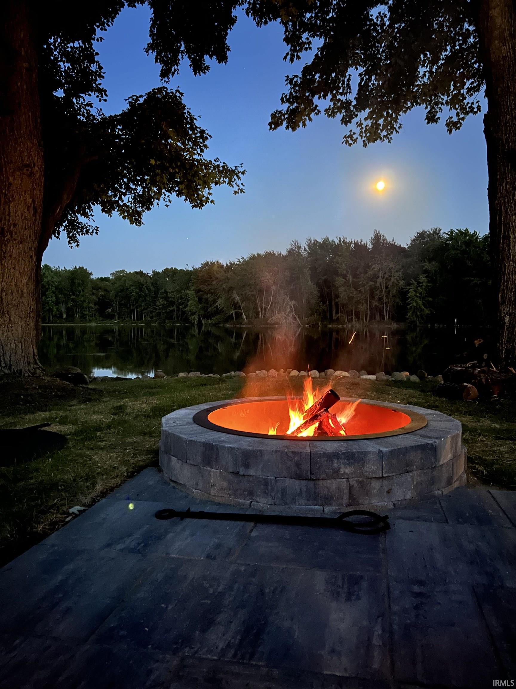 Paved patio featuring a stone fire pit, surrounded by lush green lawn and mature trees, with a body of water in the background