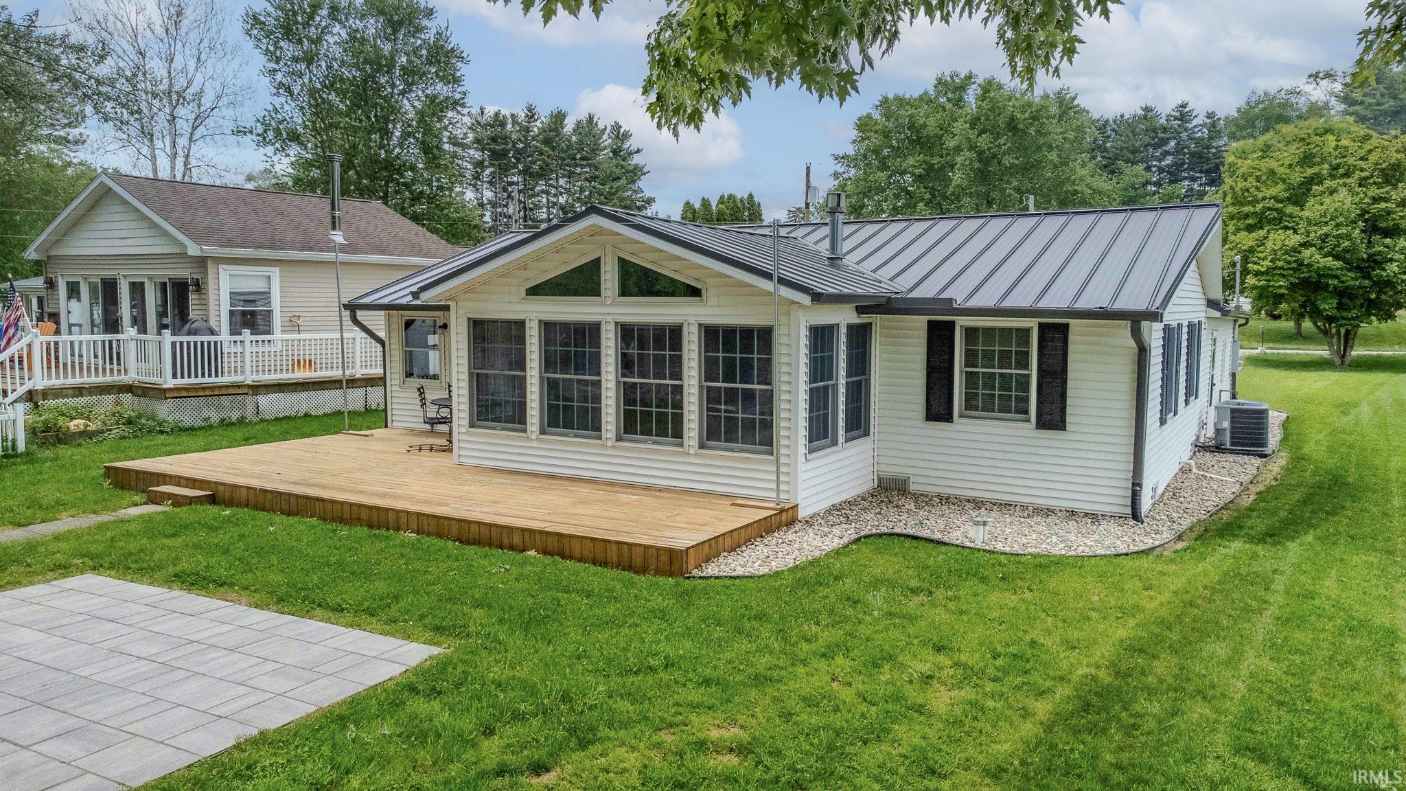 White siding exterior featuring a dark metal roof, a full-length sunroom with large windows, an expansive wooden deck, and a paver patio