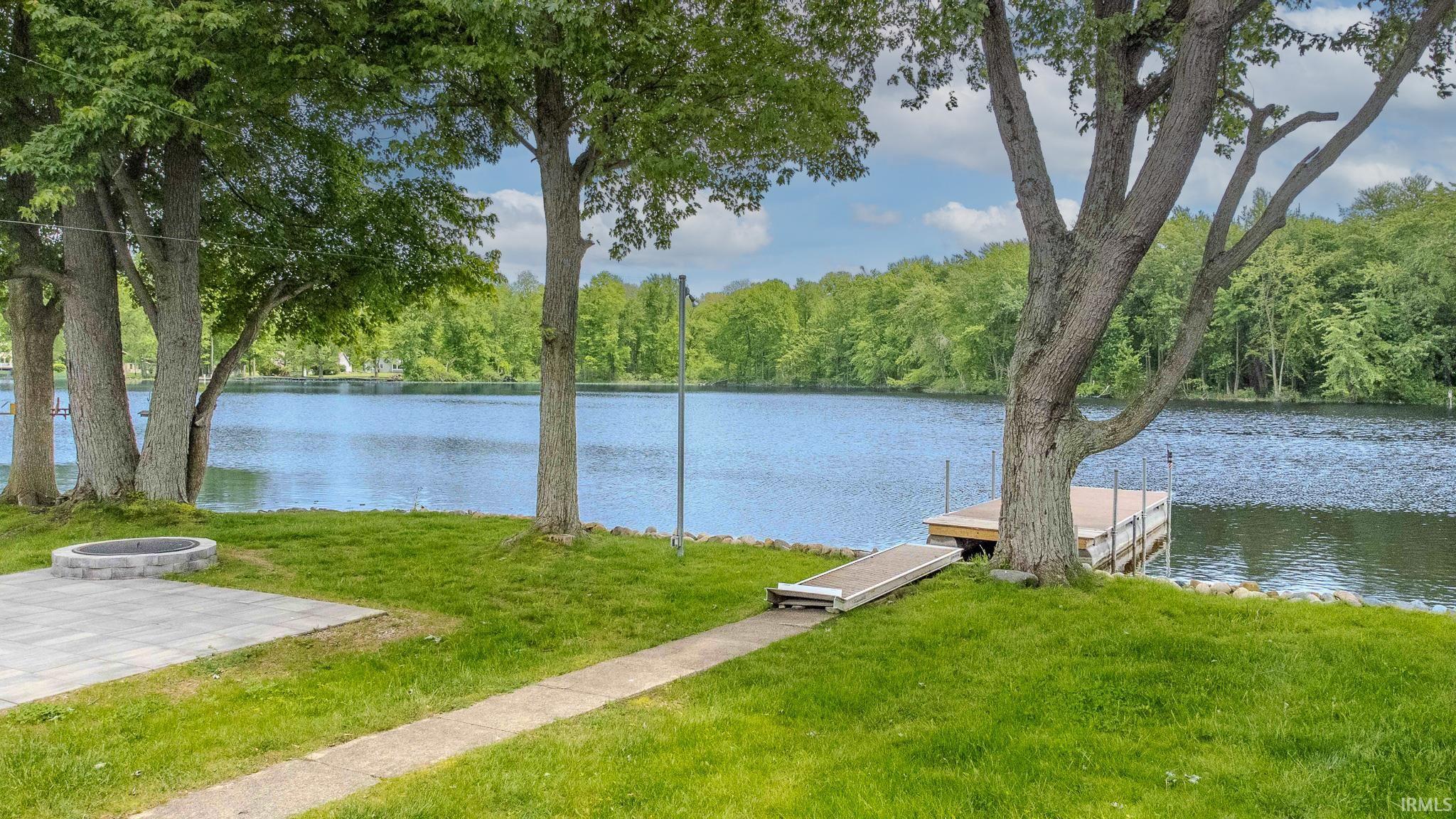 Waterfront property featuring a private dock, paved patio, built-in fire pit, mature trees, and a grassy expanse