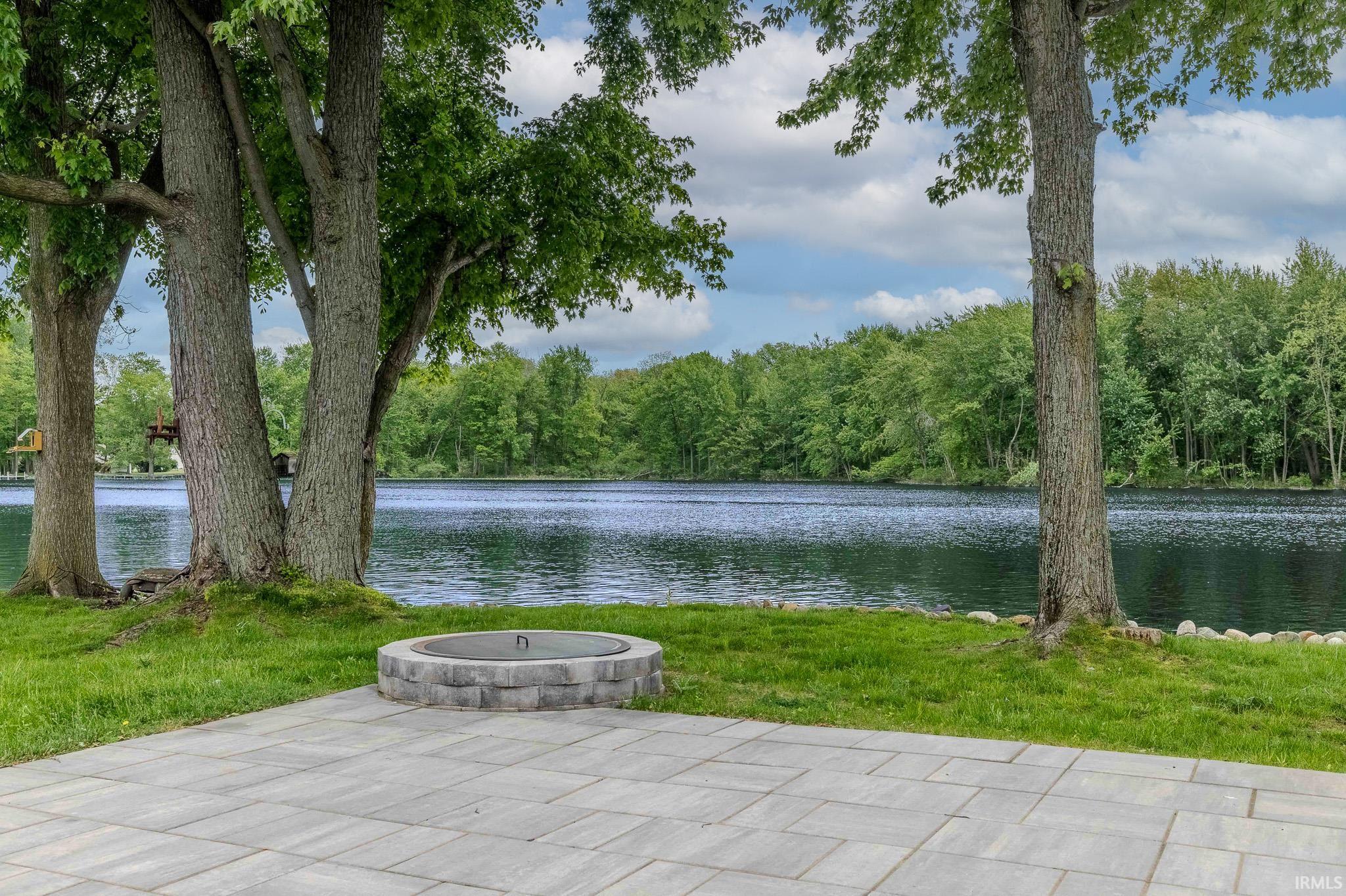 Paved patio area featuring a built-in stone fire pit, bordered by a manicured lawn and mature trees, with a serene lake backdrop
