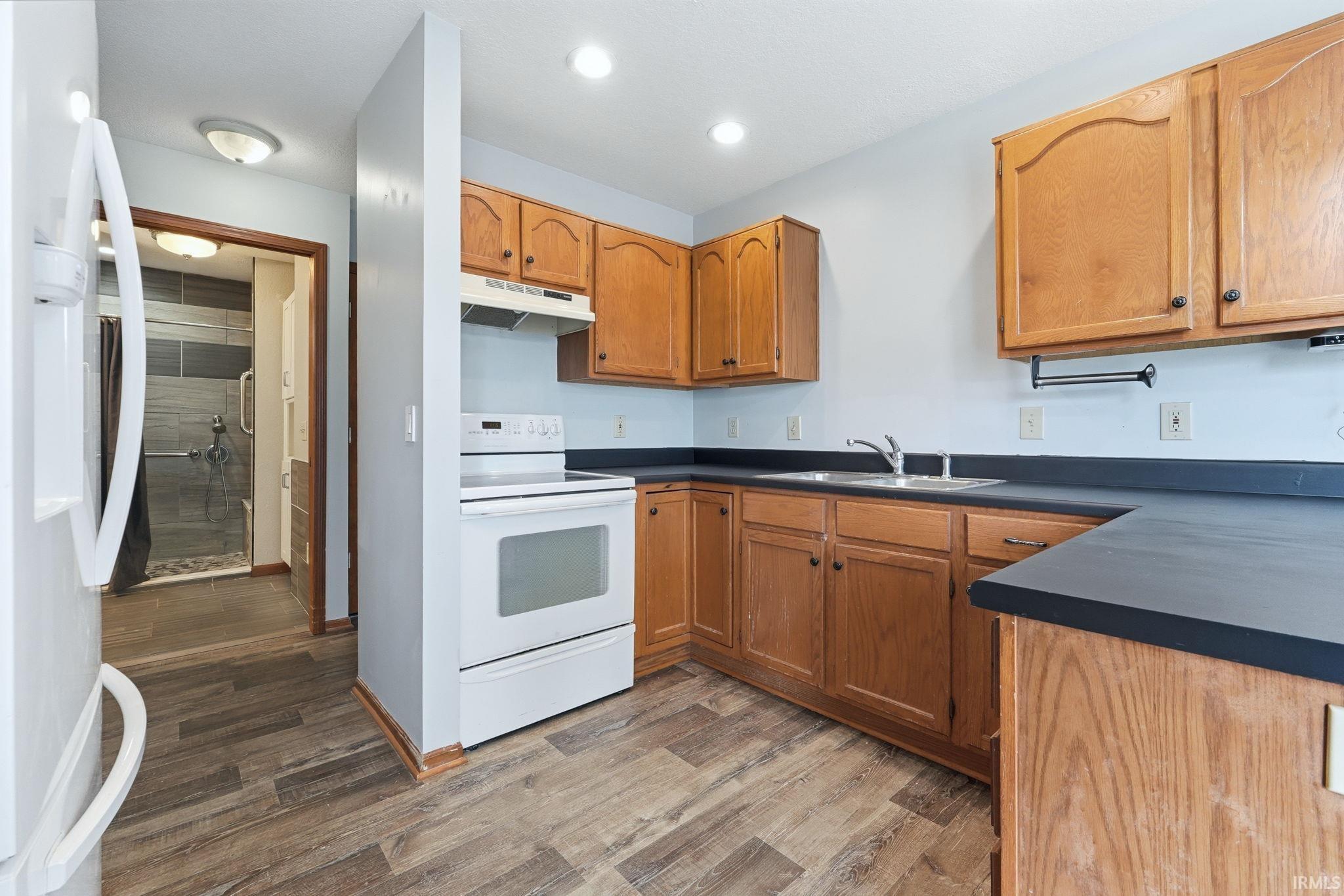 Kitchen featuring wood-finish flooring, light wood cabinetry, white appliances, and dark countertops