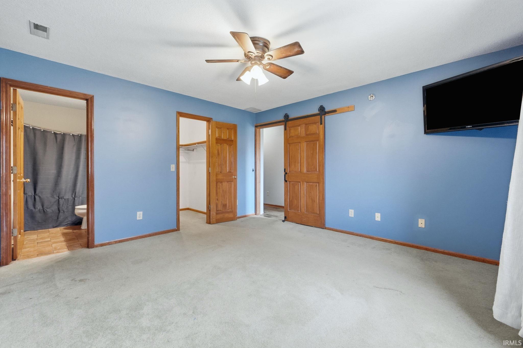 Spacious interior featuring a ceiling fan, light-toned carpeting, wood-finish trim, a barn door, and an adjacent bathroom