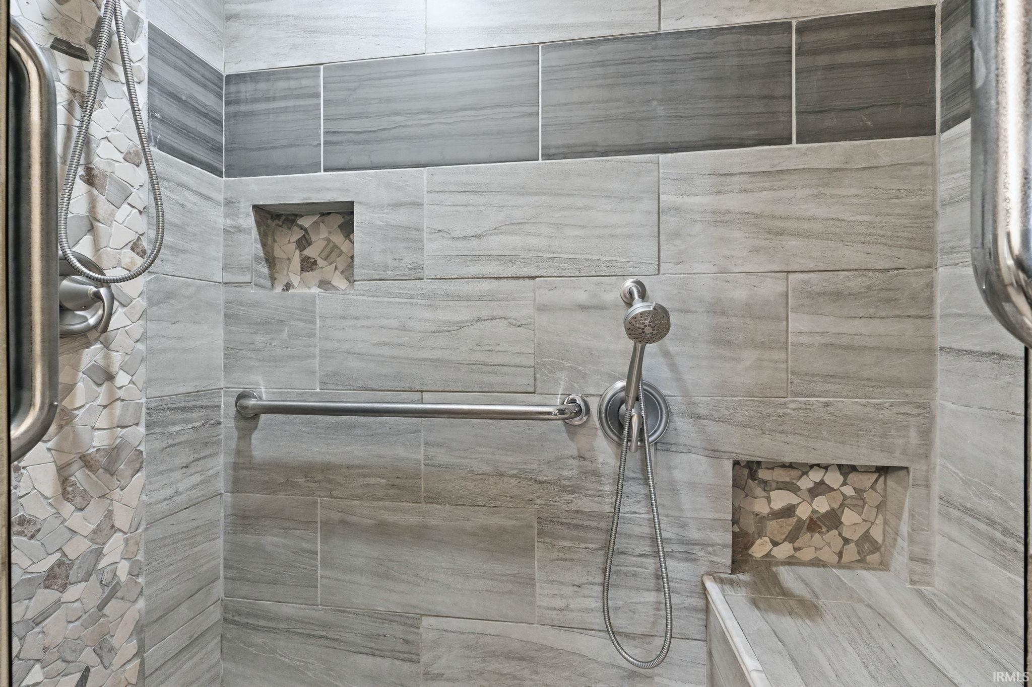 Tiled shower featuring a handheld showerhead, integrated grab bar, two recessed niches with stone mosaic accents, and a built-in bench