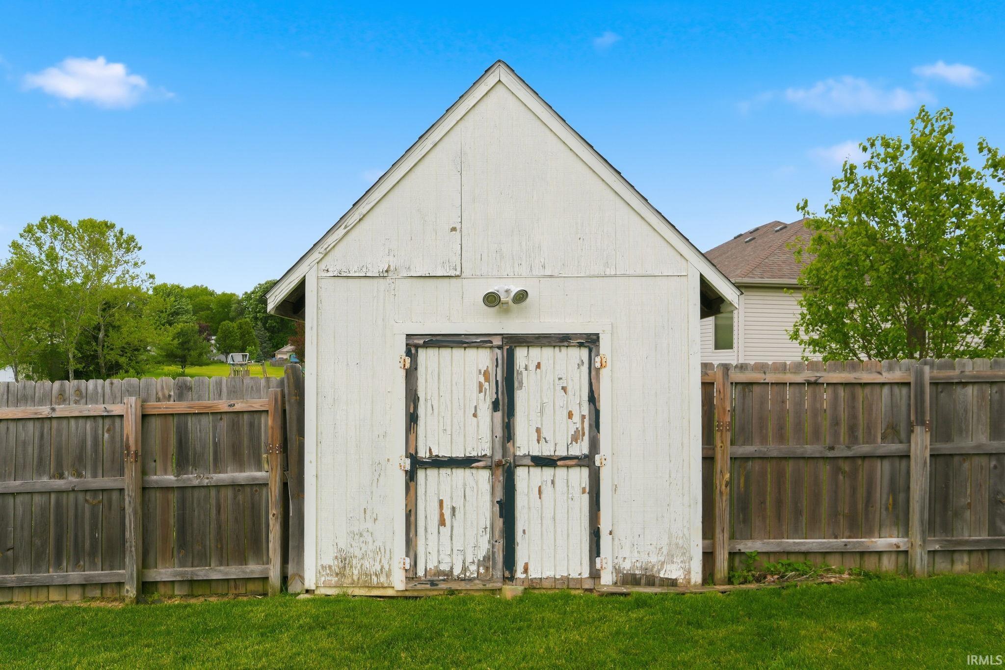 Detached storage shed with a gable roof, double doors, exterior lighting, and a surrounding wooden fence
