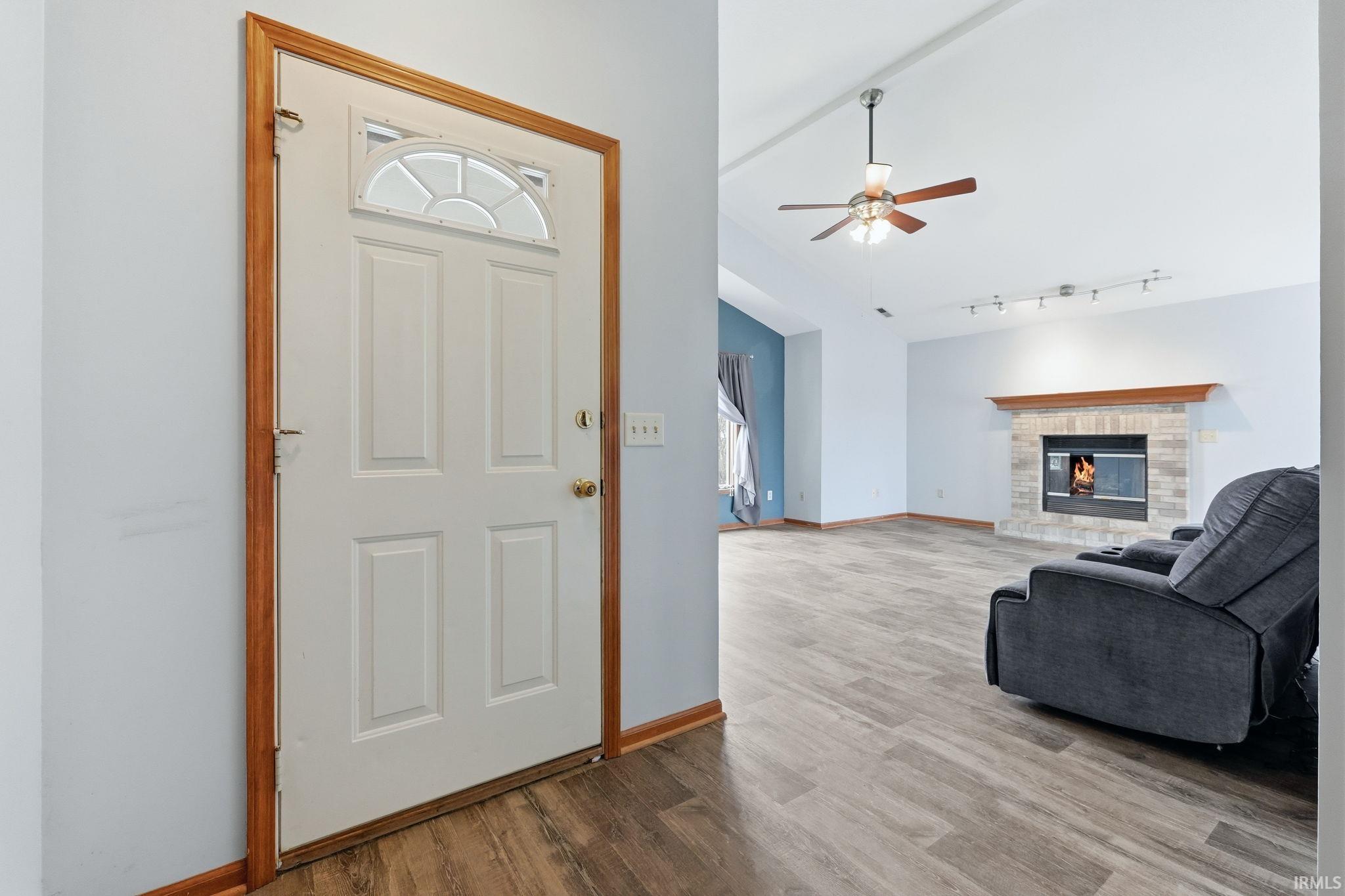 Entryway with wood-finish flooring opening to a living area featuring a vaulted ceiling with a ceiling fan, a fireplace with a brick surround and wood mantel, and track lighting