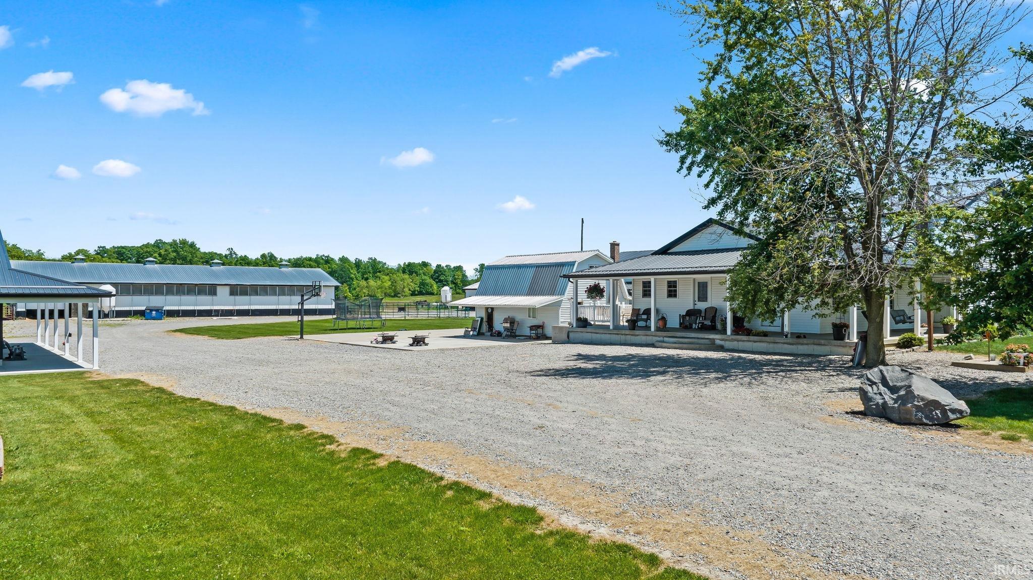 Expansive gravel driveway leading to a white-sided farmhouse with a covered front porch, accompanied by multiple outbuildings featuring metal roofs