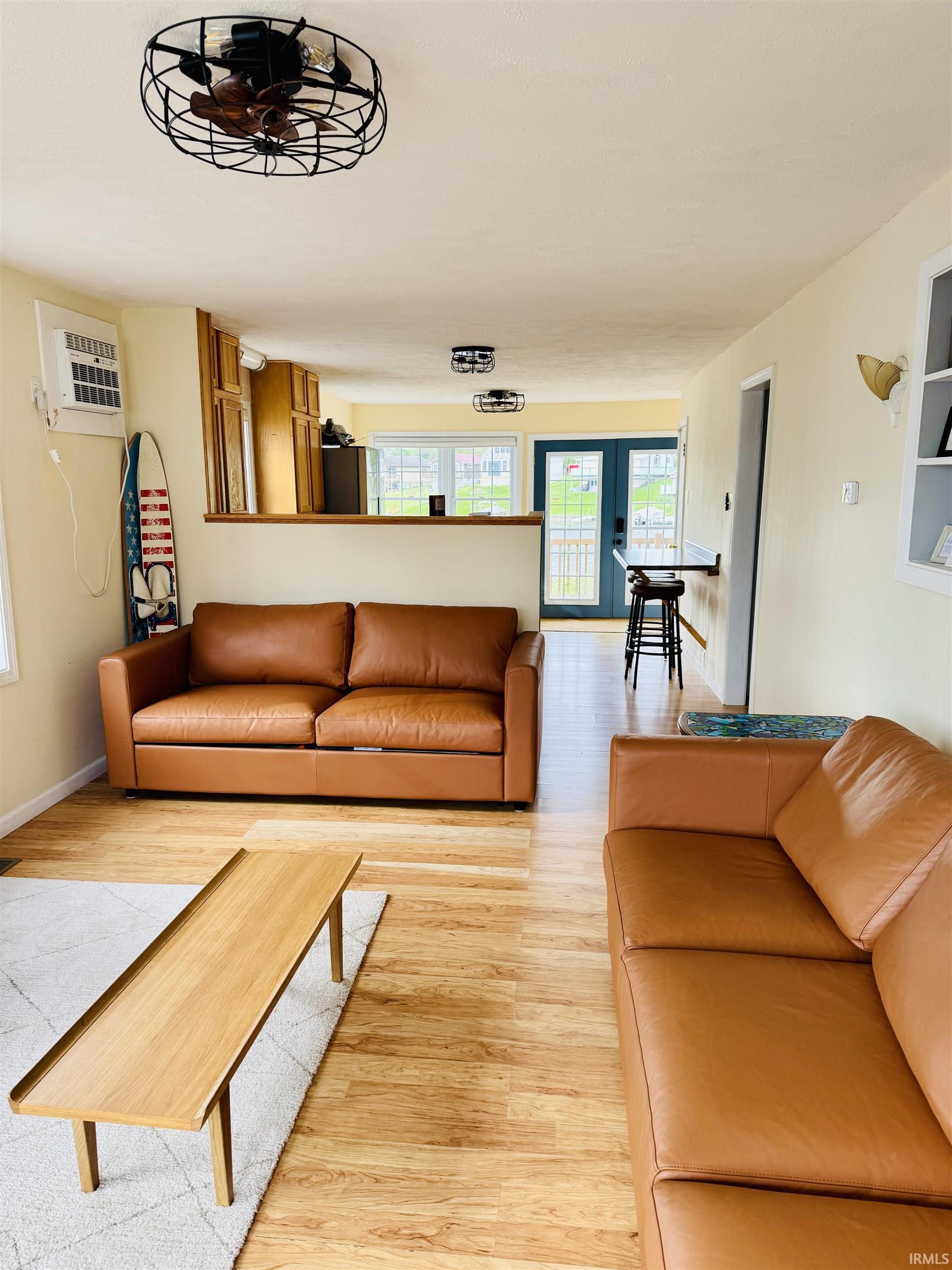 Open-concept living space with wood-finish flooring, two industrial-style ceiling fan light fixtures, a built-in air conditioning unit, and a pass-through counter to the kitchen area