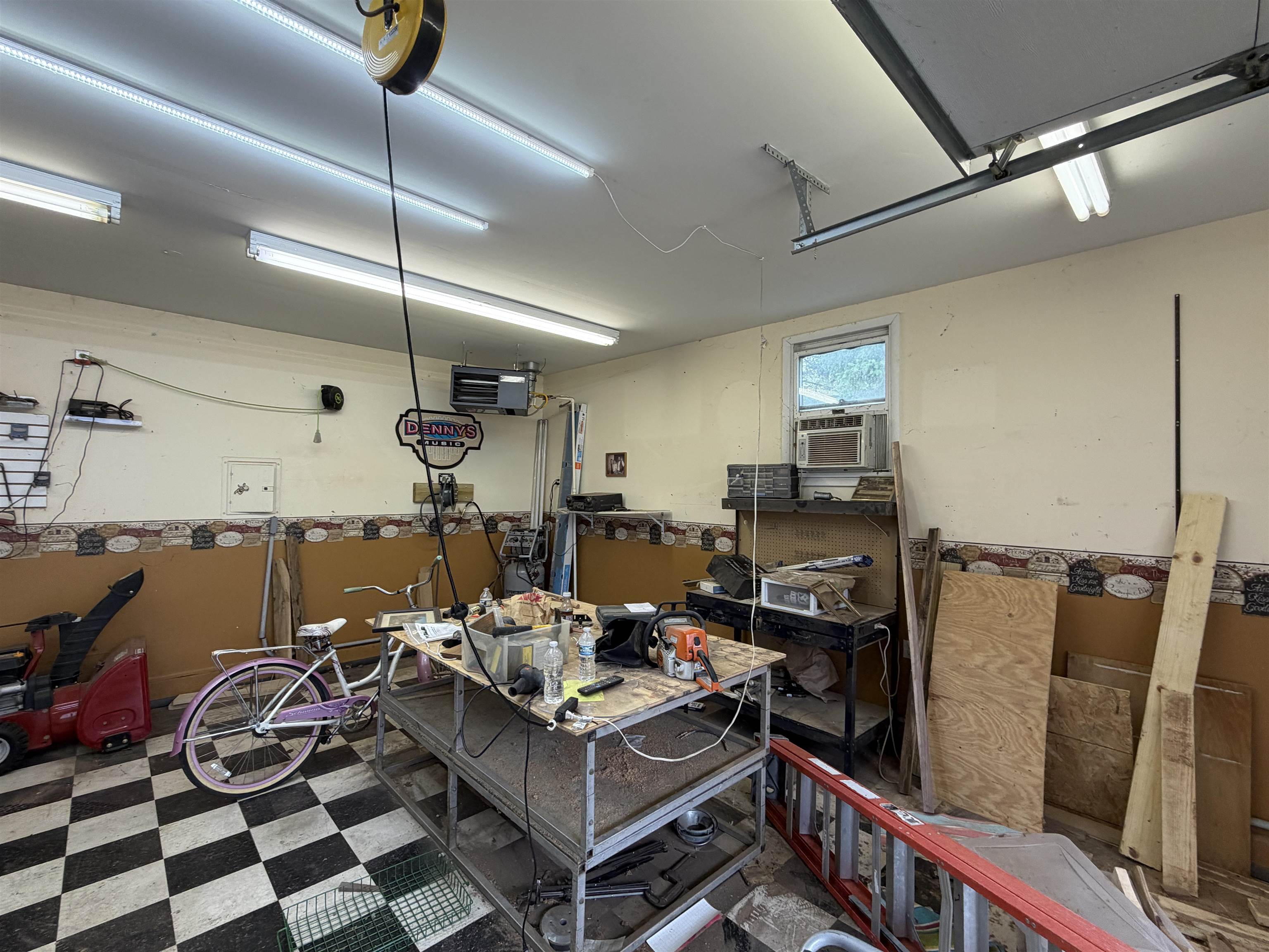 Utility room featuring checkerboard flooring, multiple fluorescent light fixtures, an exposed garage door track, a window with an air conditioning unit, and a workbench