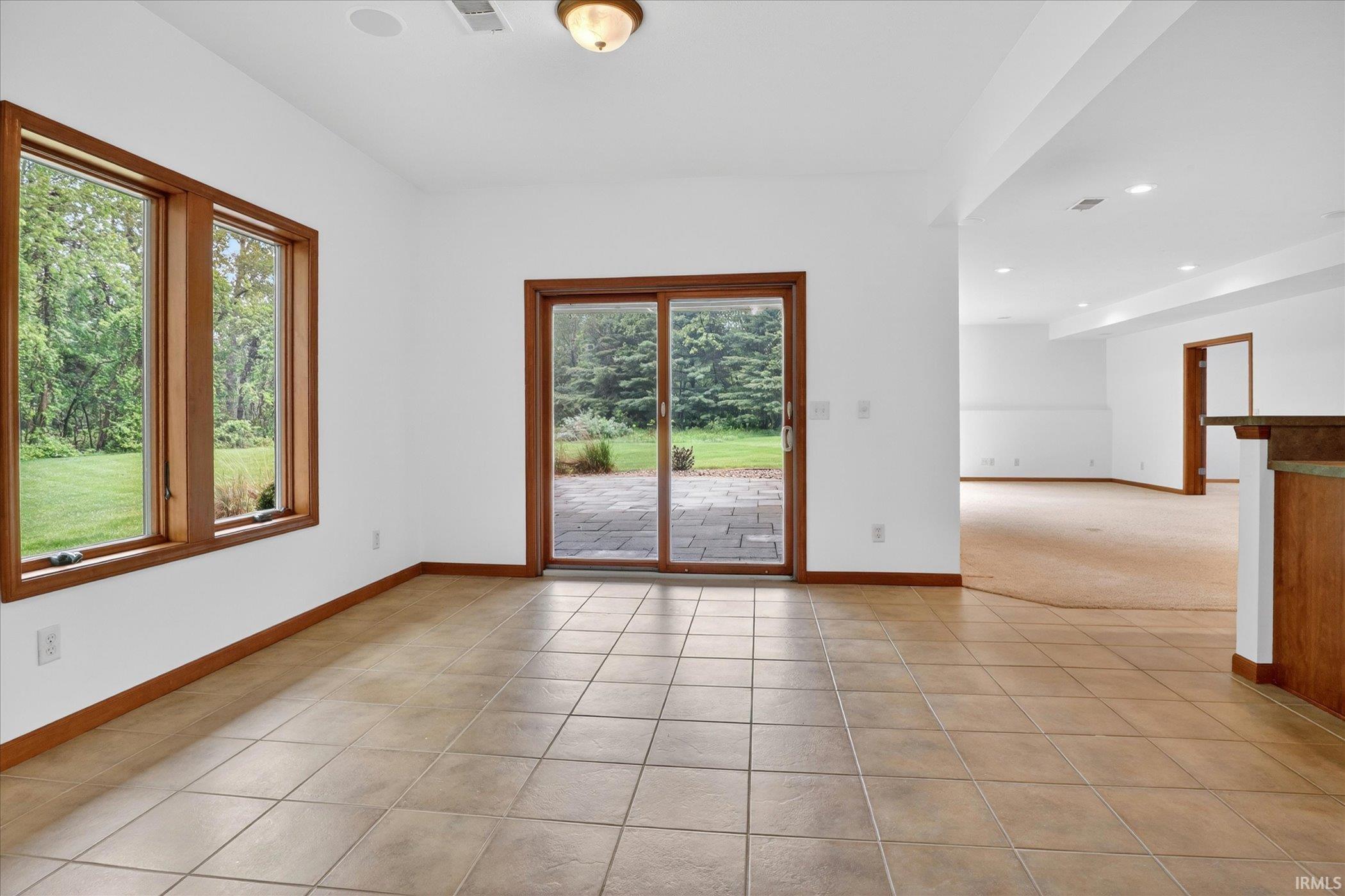 Expansive room featuring light-colored tile flooring, multiple windows with wood trim, and a glass sliding door leading to an outdoor paved patio