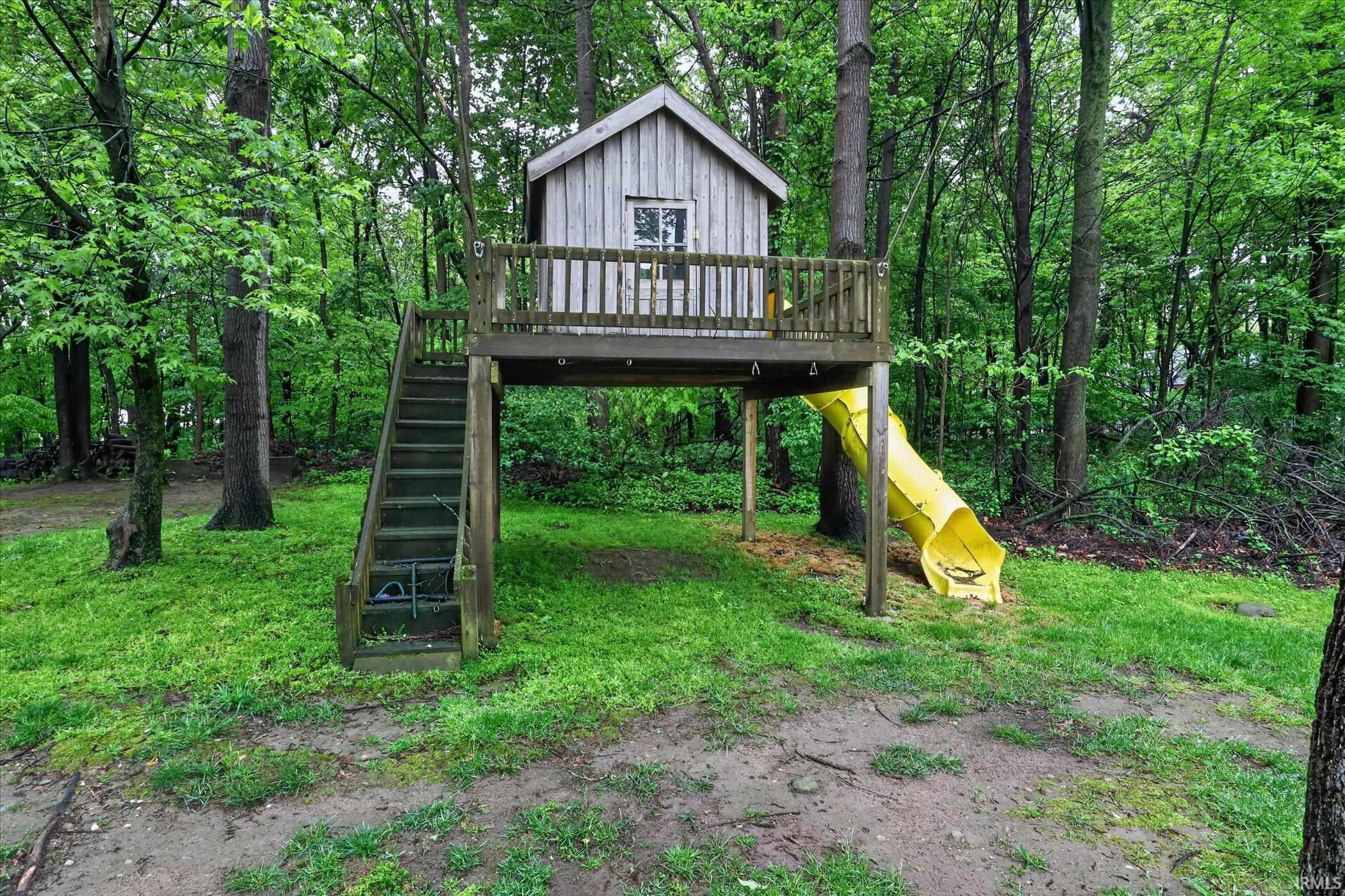 Wood-sided treehouse structure featuring an elevated deck, staircase access, and a yellow slide