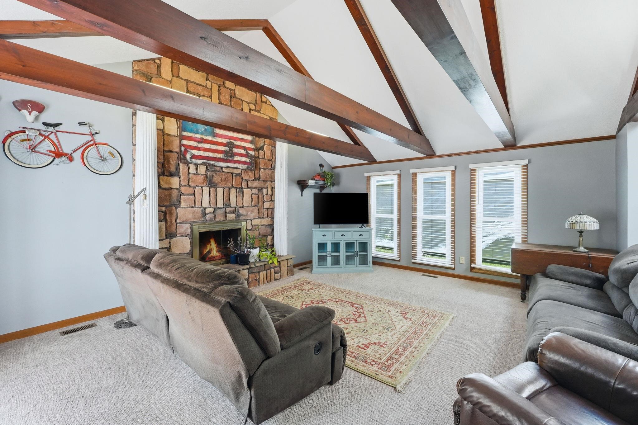 Vaulted ceiling with exposed wood beams, stone-clad fireplace, three windows with blinds, and light-toned carpeting