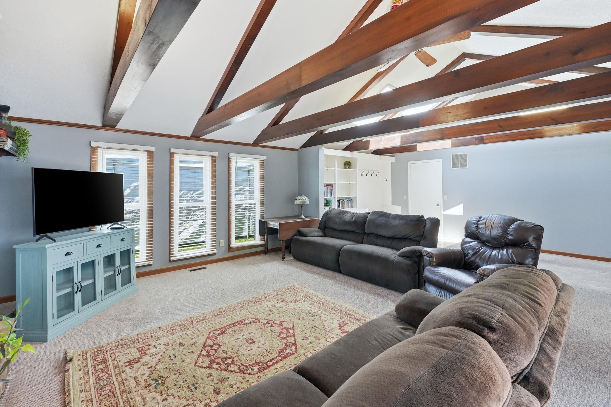 Vaulted ceiling with exposed wood beams, multiple windows, carpeted flooring, and built-in shelving