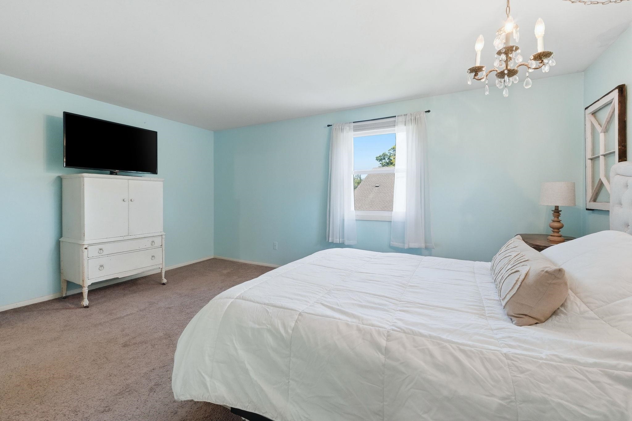 Bedroom featuring light blue walls, carpeted flooring, a window with white sheers, and a chandelier with decorative crystals
