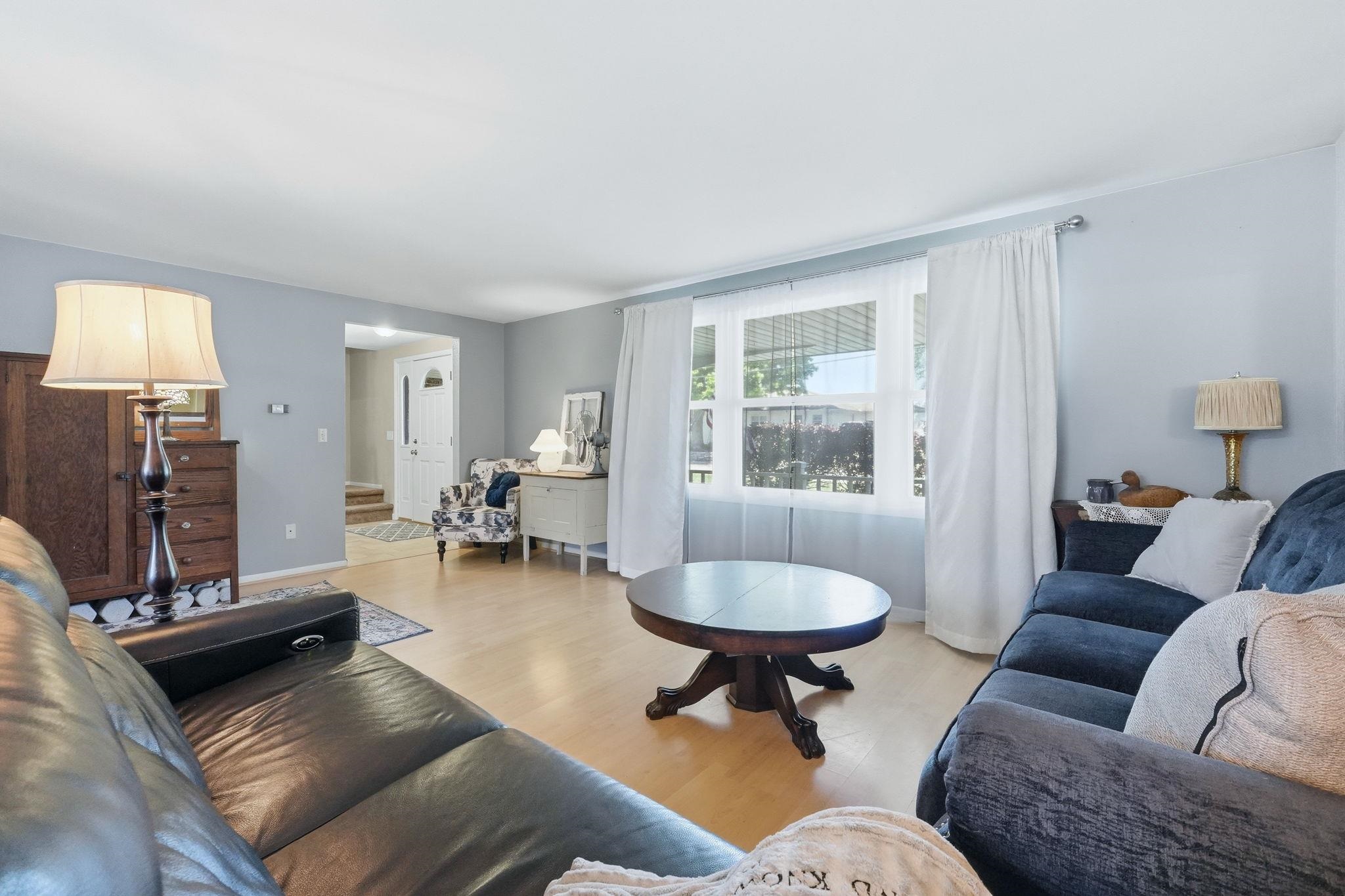 Spacious living area featuring light wood-finish flooring, a large window with white curtains, and light gray wall paint