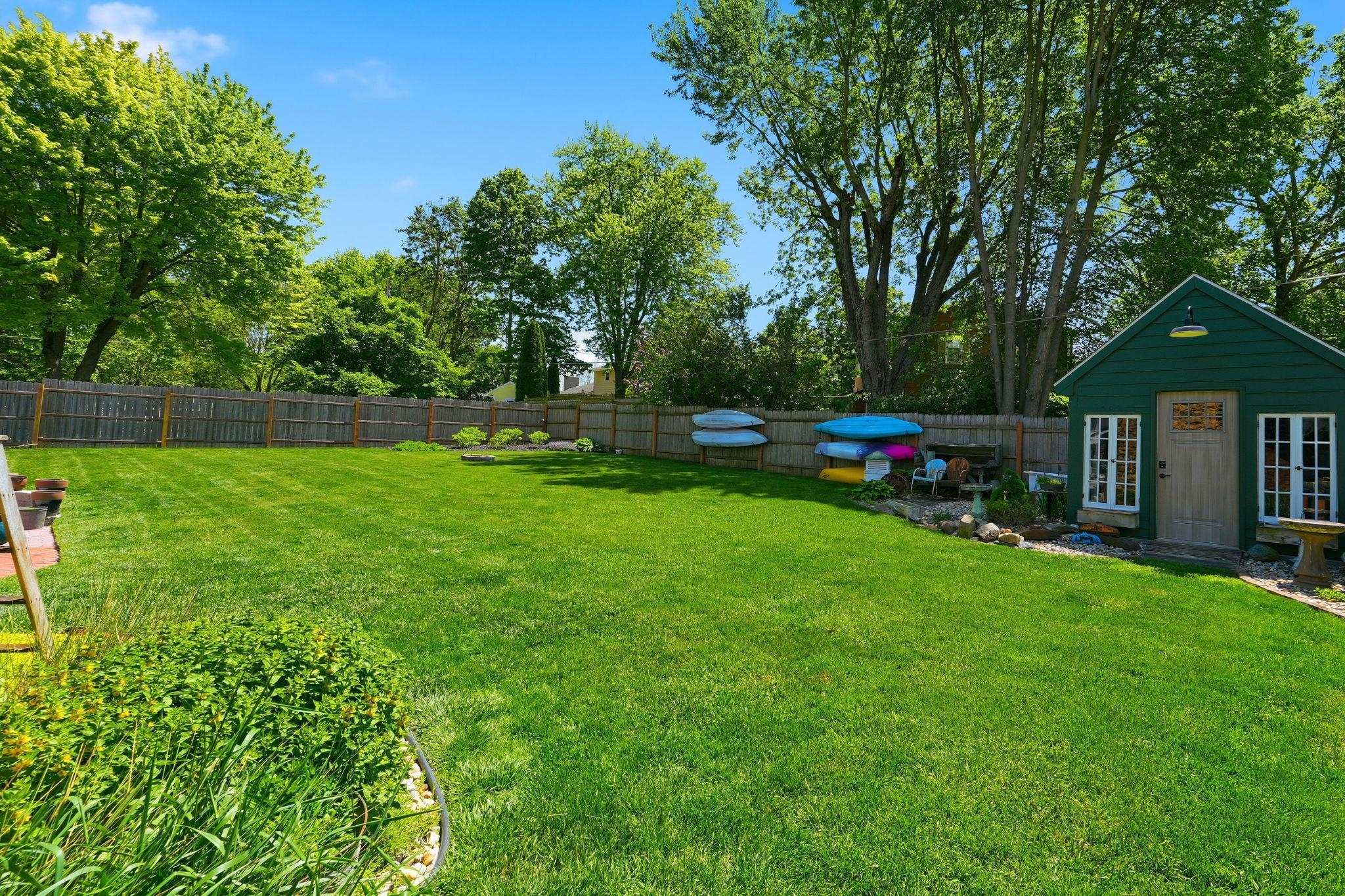 Expansive backyard featuring a manicured lawn, full perimeter wood fencing, mature trees, a green storage shed with multiple windows, and defined landscape beds