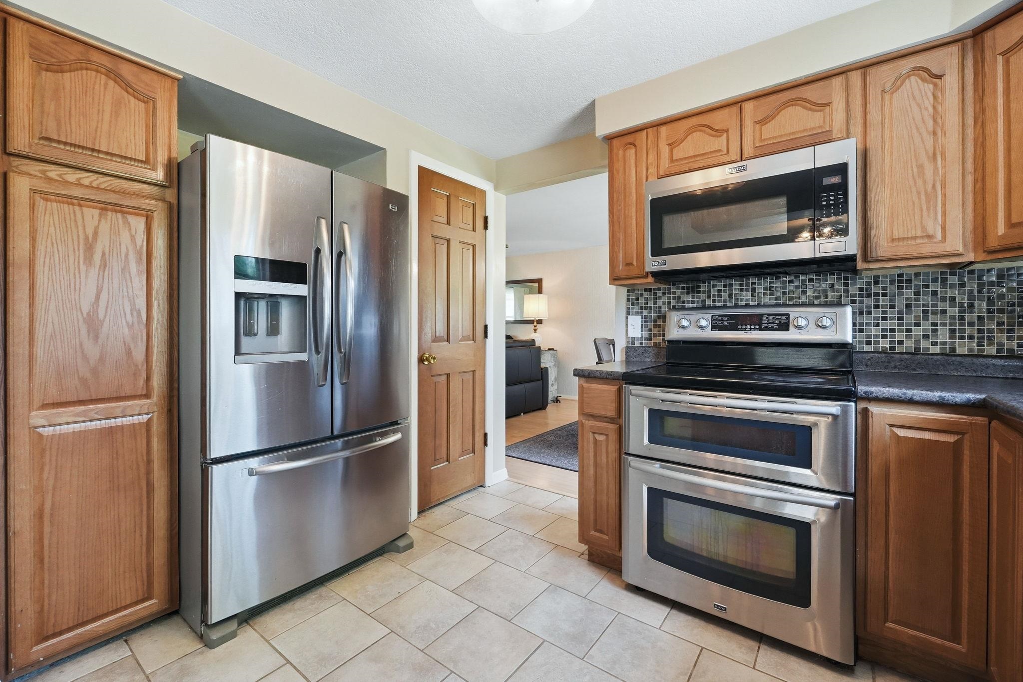 Kitchen featuring wood cabinetry, stainless steel appliances, a black countertop, a mosaic tile backsplash, and light-toned tile flooring