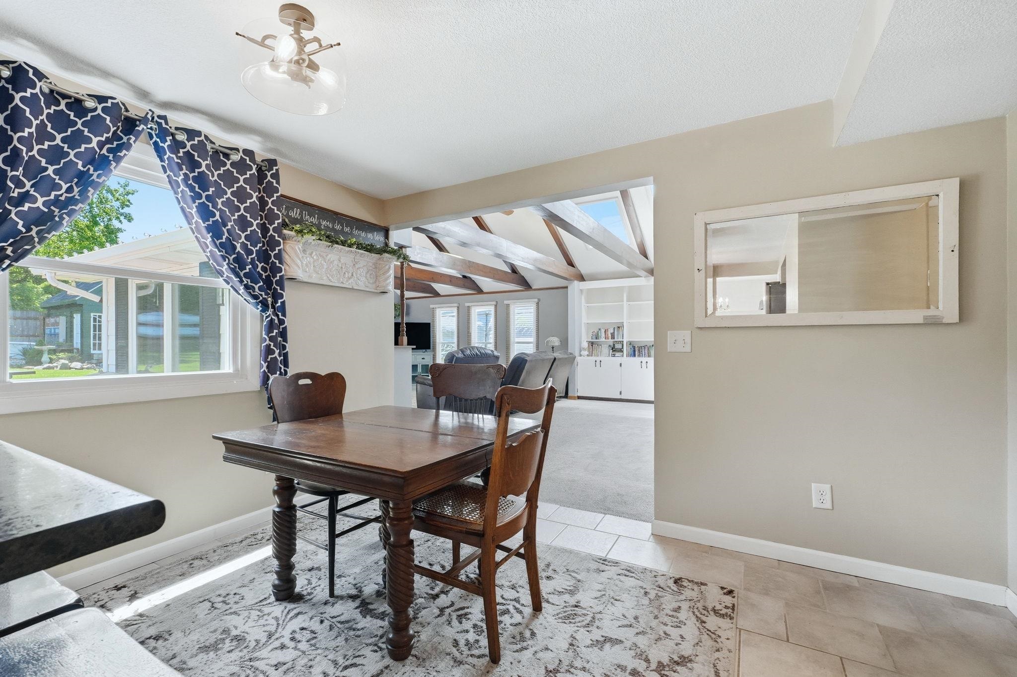 Dining space featuring a window with exterior views, tile flooring, and an open doorway to an additional room with exposed ceiling beams and built-in shelving