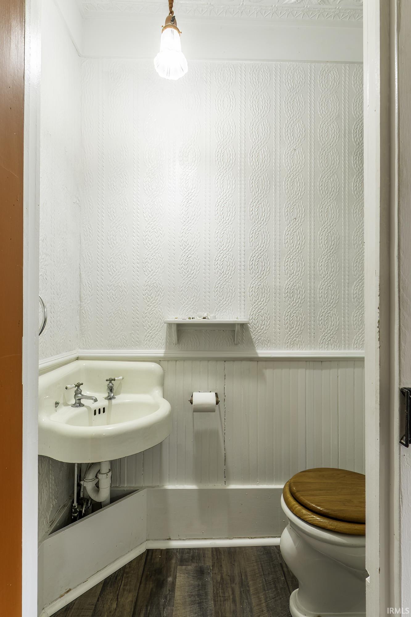 Compact powder room featuring wood-finish flooring, a corner-mounted sink, a toilet with a wooden seat, beadboard wainscoting, and textured wallpaper