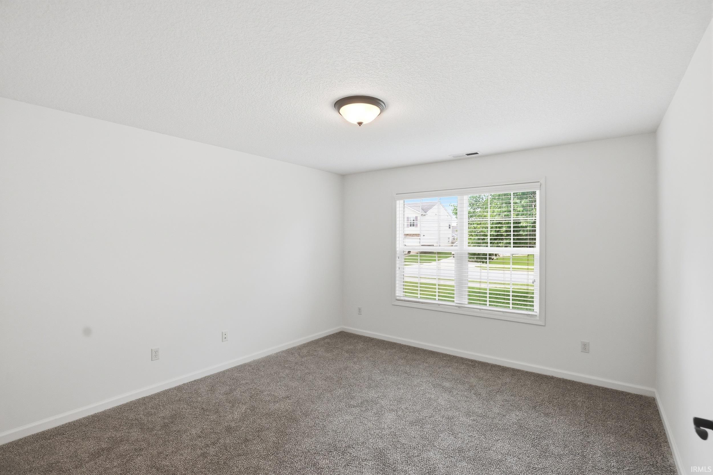 Spacious room featuring neutral carpeting, white baseboards, a flush-mount ceiling light fixture, and a large window with white blinds
