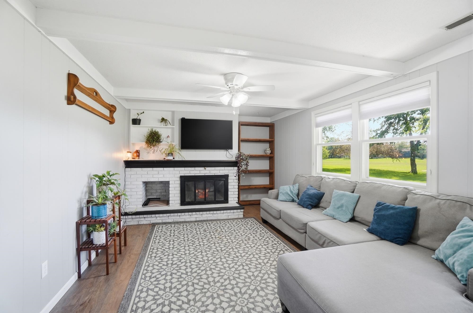 Living area featuring a white brick fireplace with a black mantel, built-in shelving, wood-finish flooring, and white ceiling beams