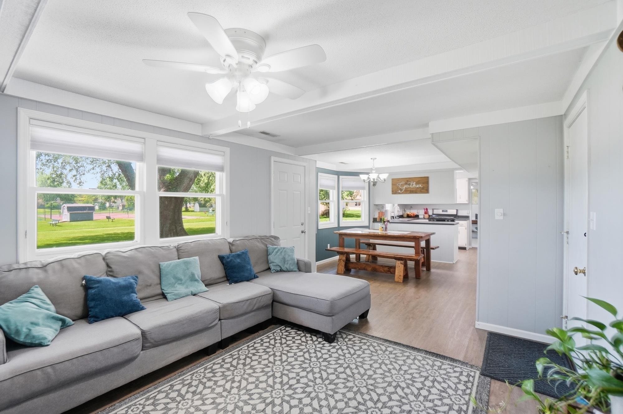 Open-concept living area featuring a ceiling fan with integrated lighting, multiple windows, and wood-finish flooring extending into the kitchen
