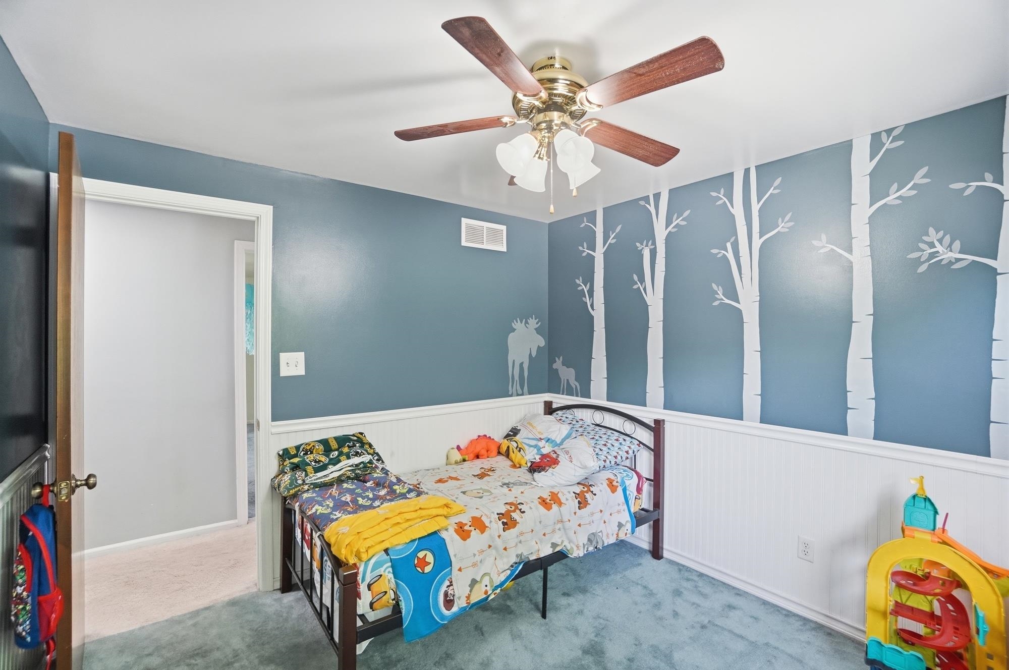 Room featuring a ceiling fan with light fixture, painted walls, wall art, wainscoting, and carpet flooring