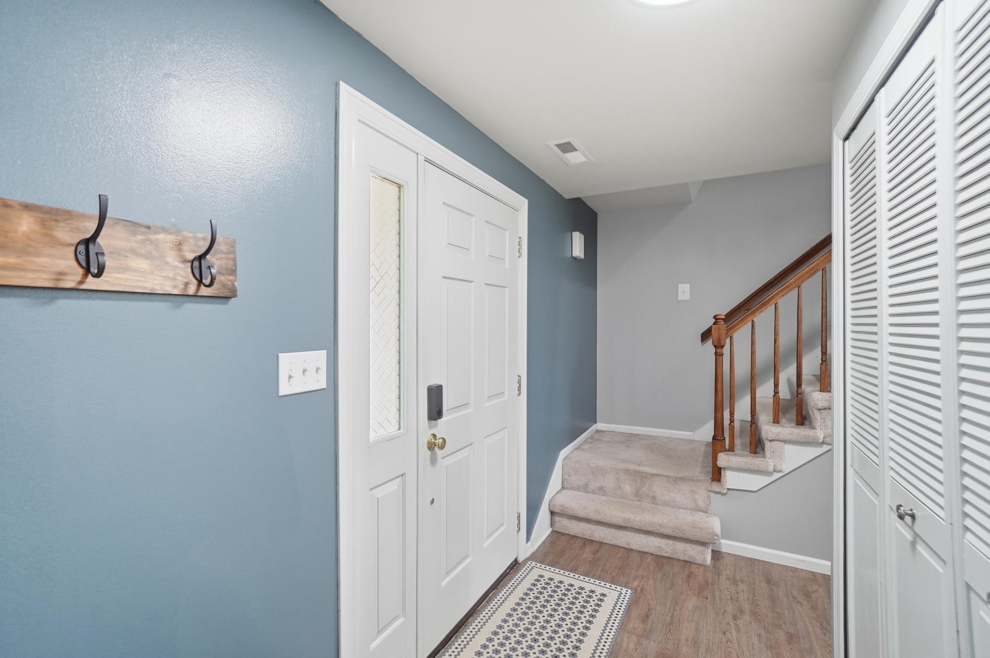 Entryway featuring a white paneled door with glass insert, wood-finish flooring, a carpeted staircase with wood handrail, and white louvered bifold closet doors