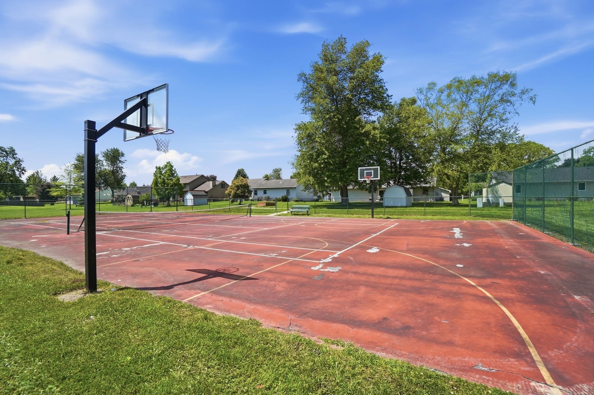 Community basketball court featuring multiple hoops, painted court lines, a surrounding green chain-link fence, and mature trees