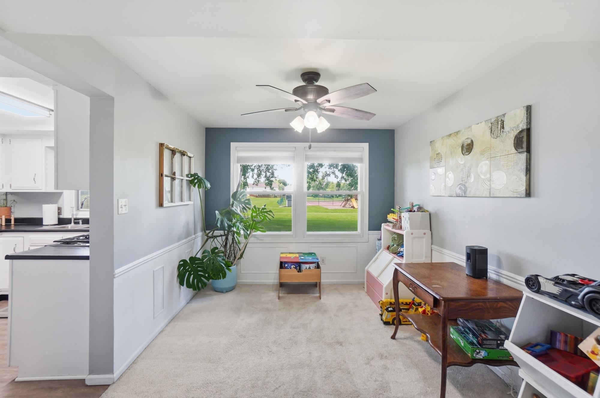 Carpeted room with wainscoting and a ceiling fan, featuring a window overlooking a grassy outdoor area