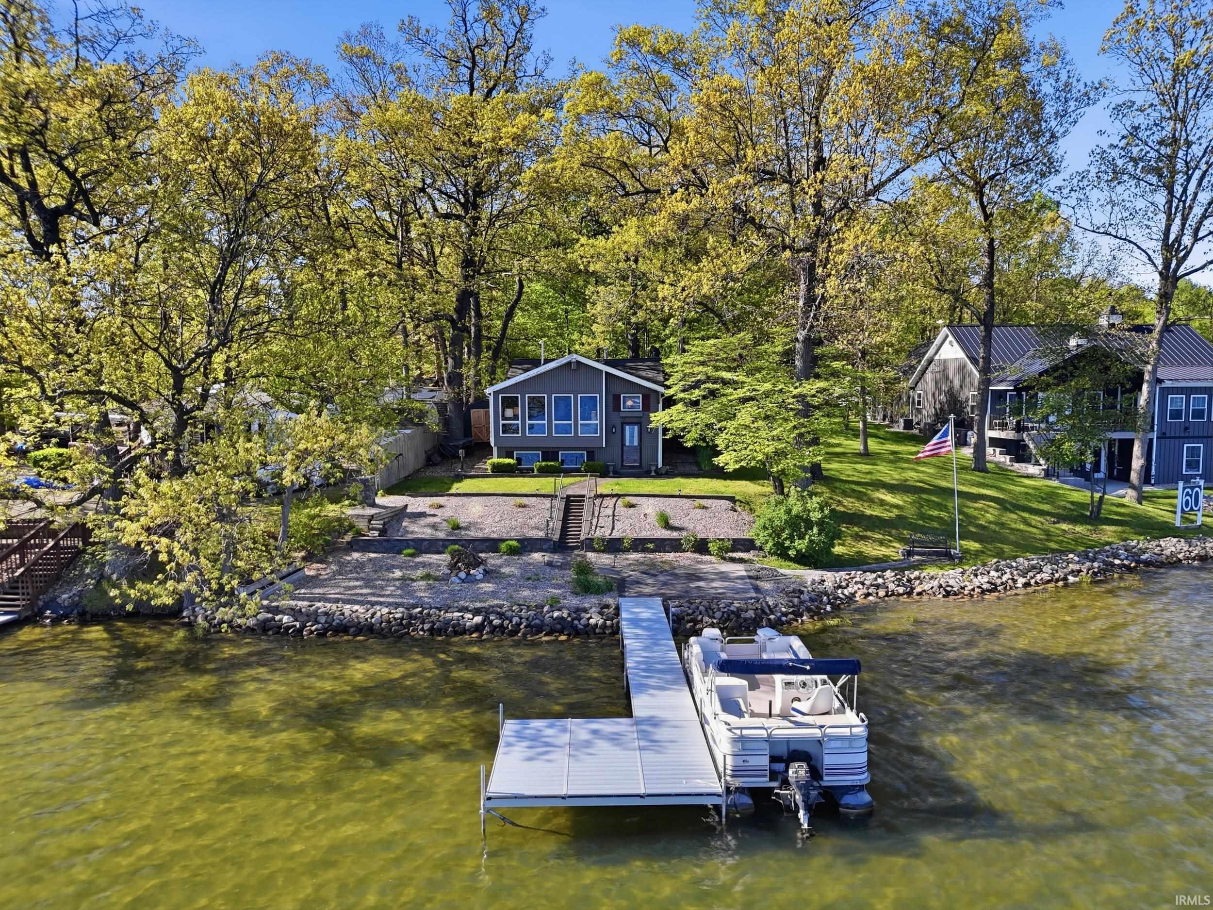 Waterfront property featuring a private dock, rocky shoreline, mature trees, and a tiered lawn