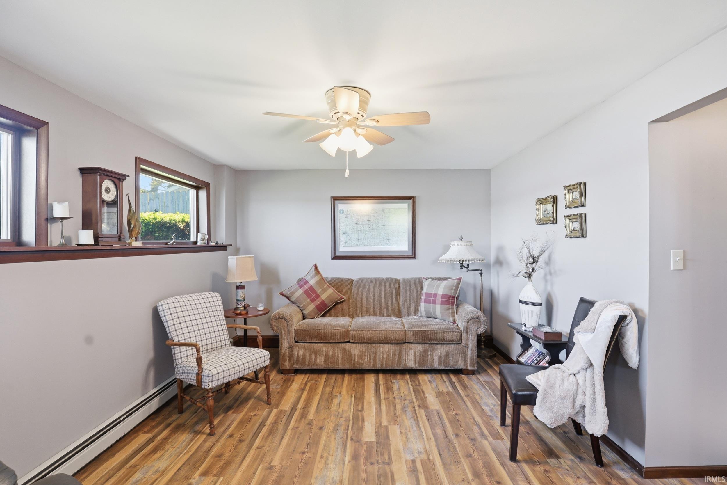 Living area featuring wood-finish flooring, light gray walls, a ceiling fan with integrated lighting, and multiple windows with dark wood trim