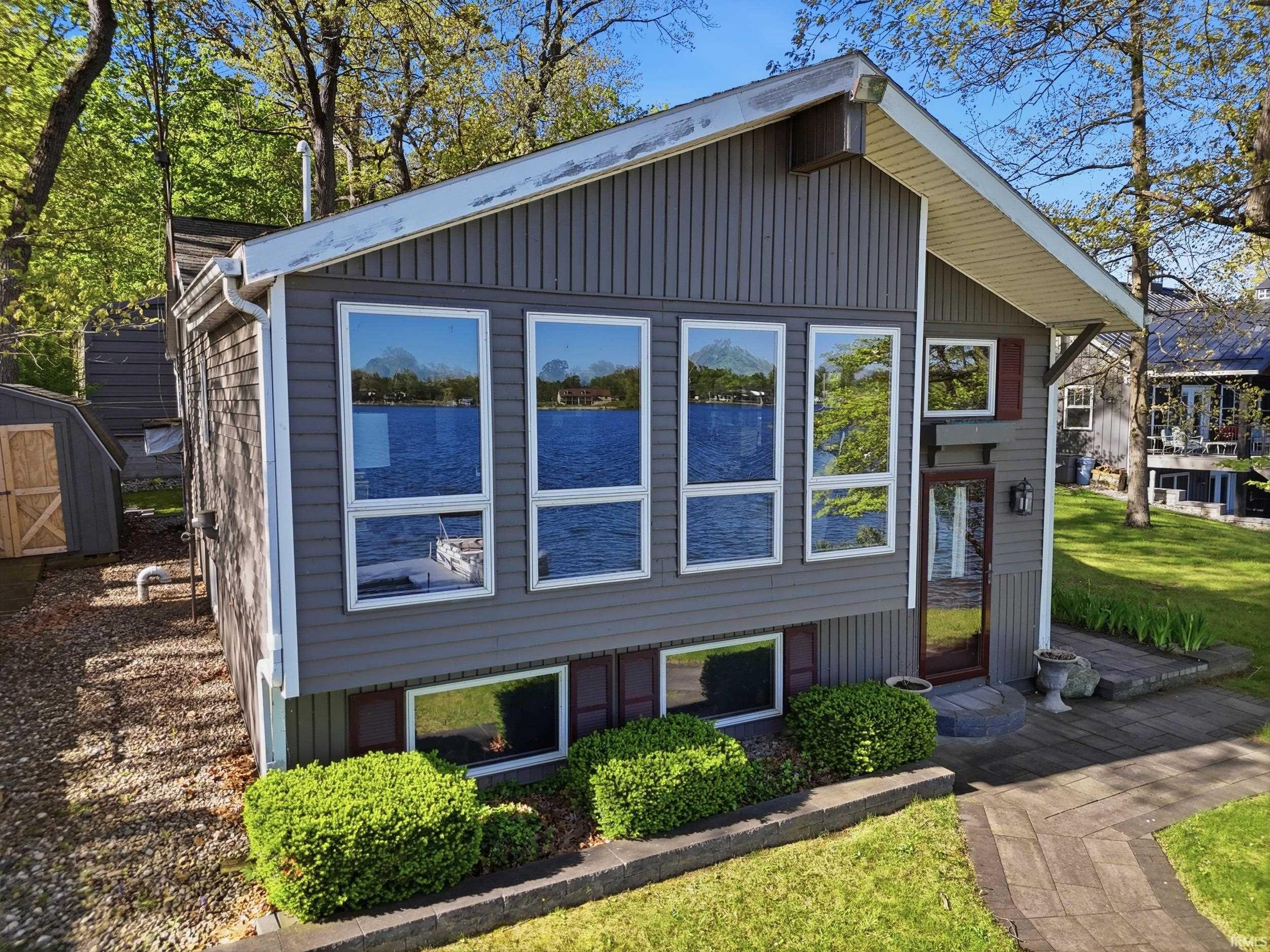 Gray vertical siding exterior featuring multiple large windows, a prominent glass-paneled entry door, mature trees, and a stone paver walkway