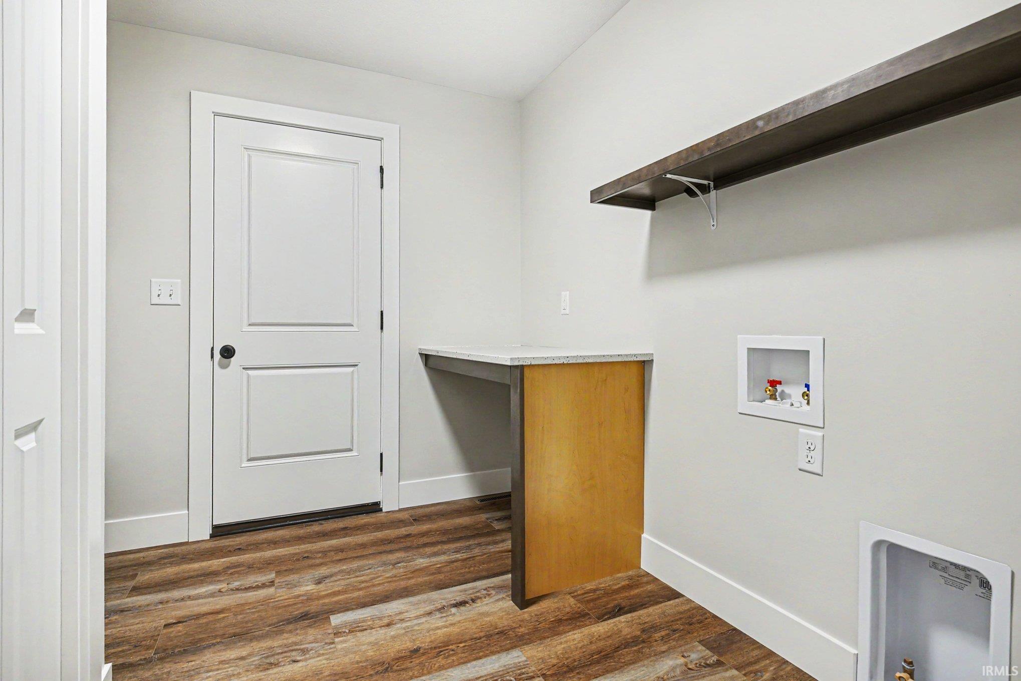 Laundry area featuring wood-finish flooring, a built-in countertop with a light-toned surface, a dark wood-finish wall shelf, and white baseboards