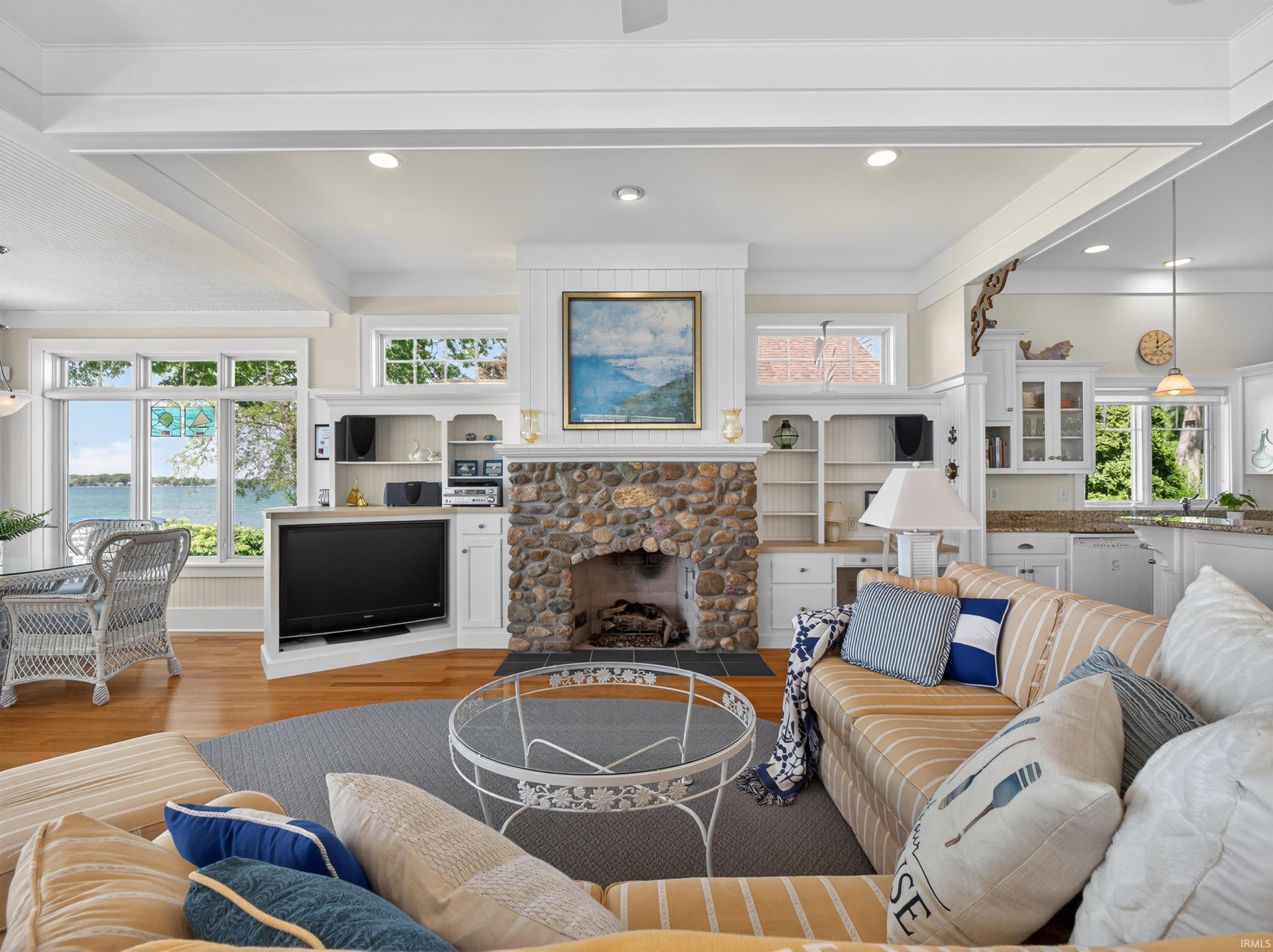 Spacious living area featuring a prominent stone-clad fireplace, hardwood flooring, built-in shelving, white coffered ceilings, and expansive windows offering clear water views
