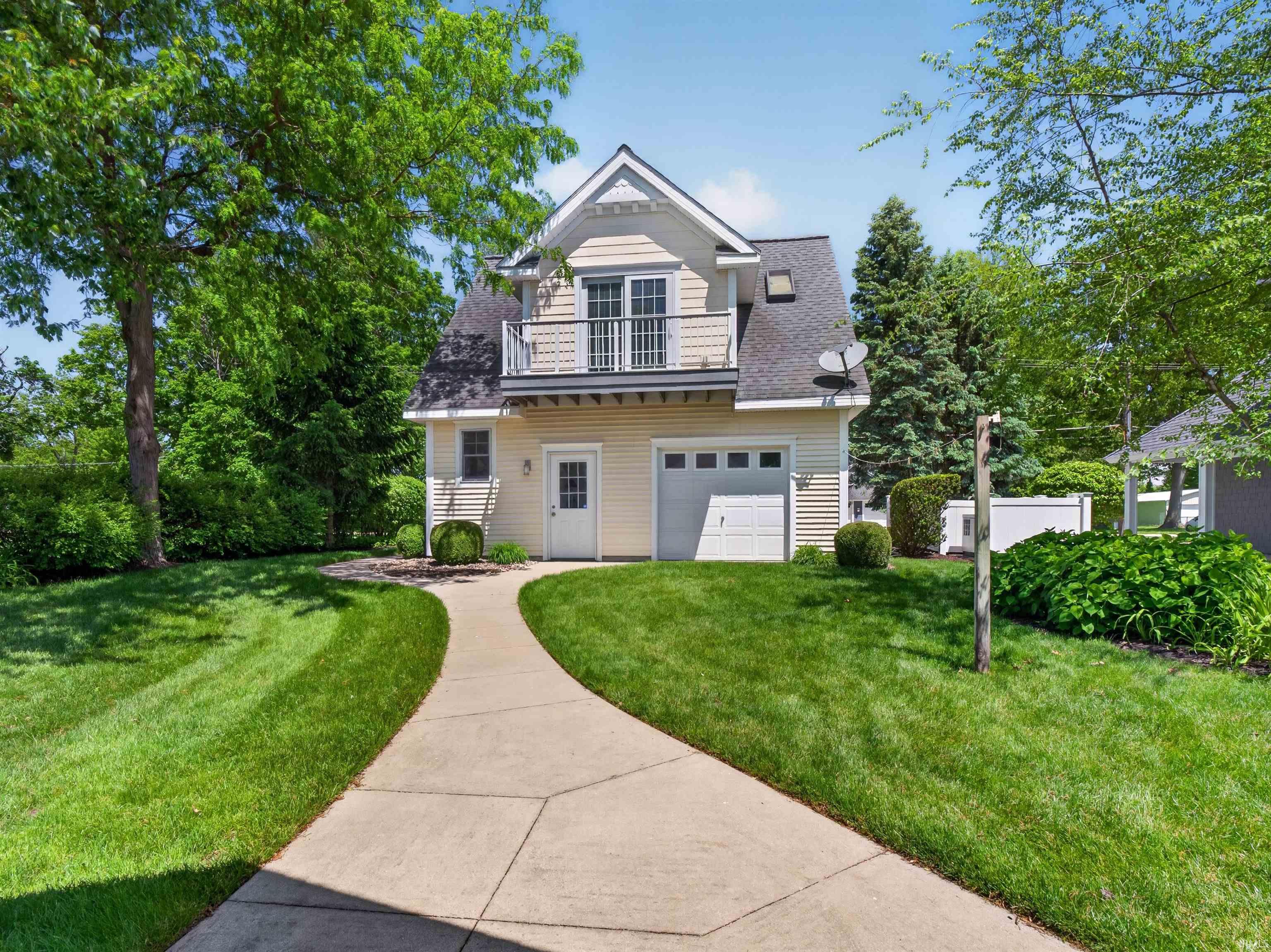 Two-story residence with light-toned siding, a second-story balcony with railings, an attached single-car garage, mature trees, and a concrete pathway