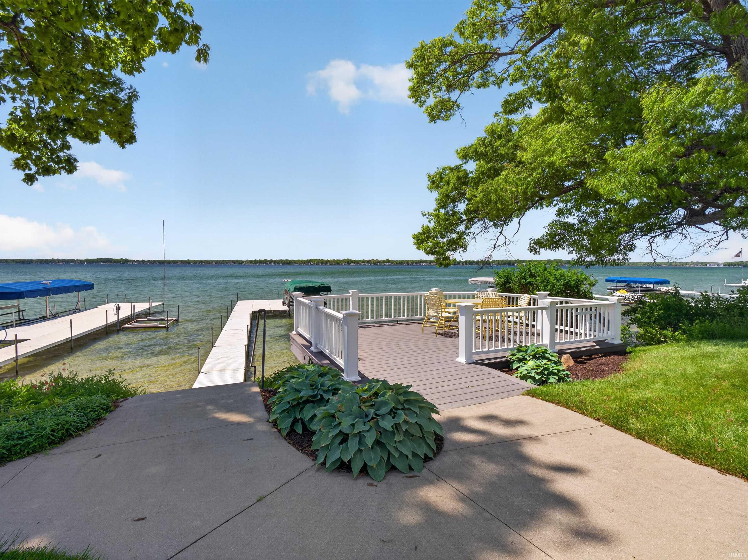 Expansive waterfront property featuring a composite deck with white railings, direct access to multiple docks, a concrete pathway, and mature trees providing natural shade