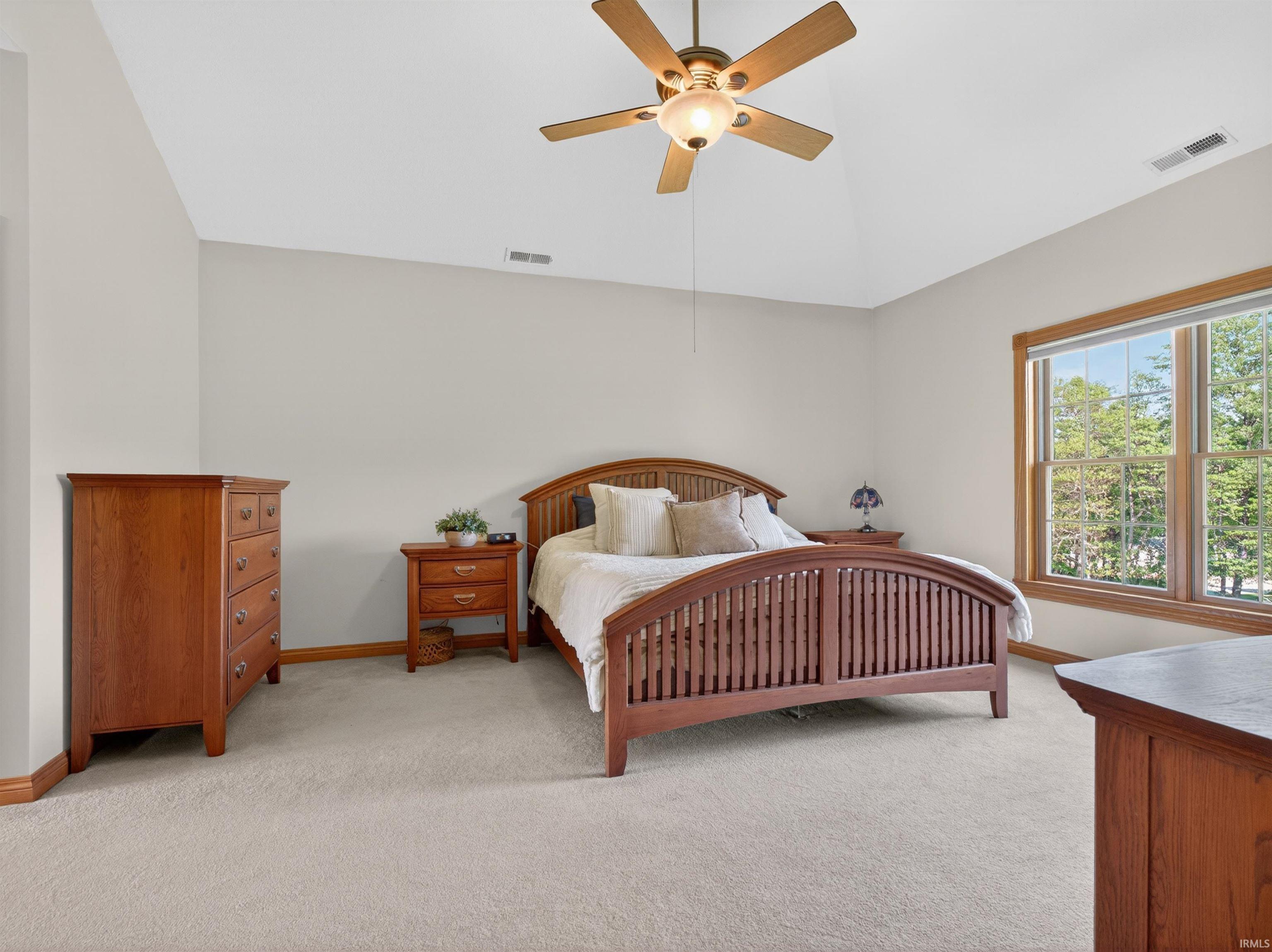 Spacious room featuring vaulted ceilings, a ceiling fan with integrated lighting, expansive window with wooden trim, light-toned carpeting, and neutral wall paint