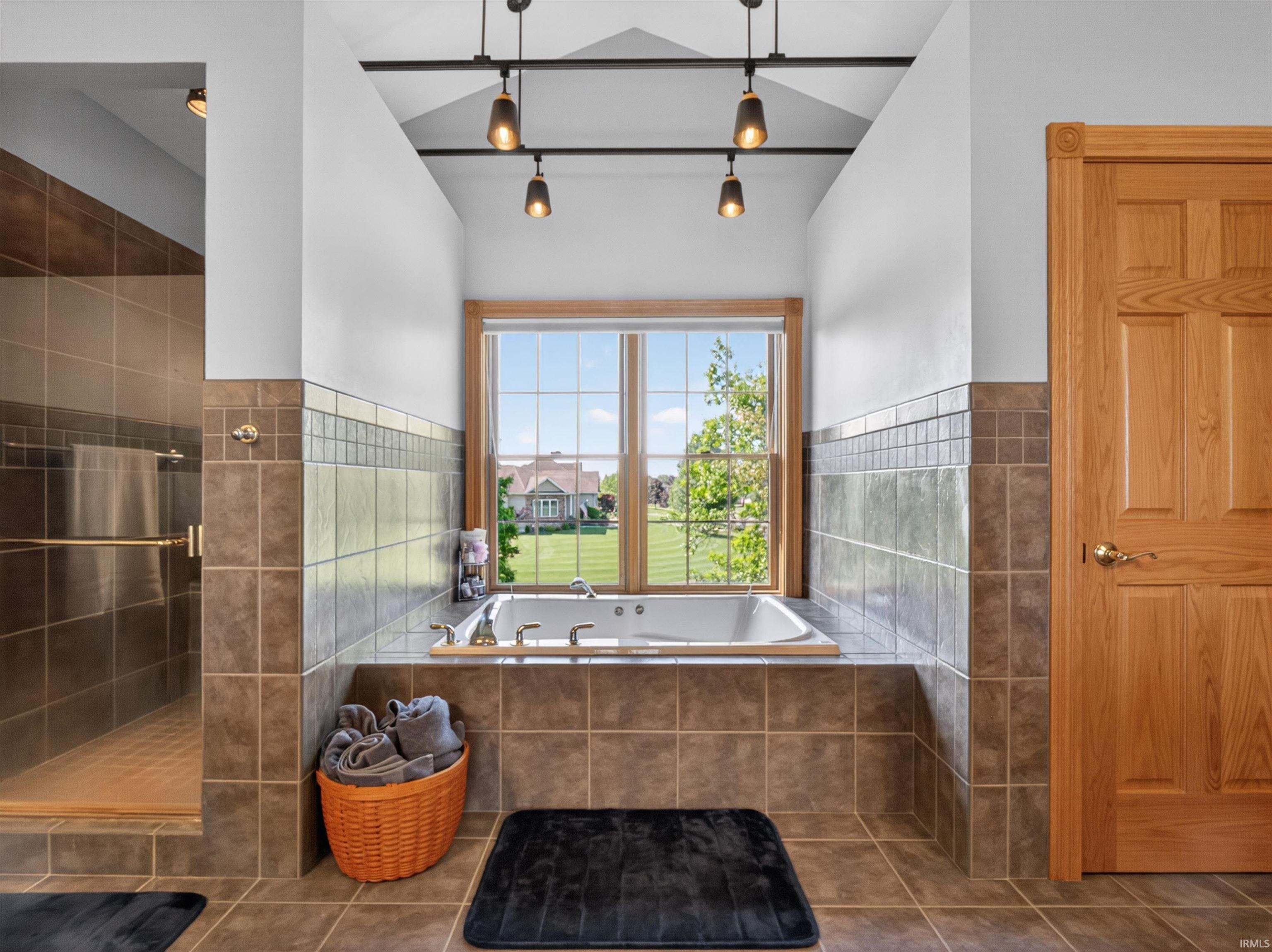 Bathroom featuring a built-in jetted tub under a large window, fully tiled shower with glass enclosure, and vaulted ceiling with track lighting