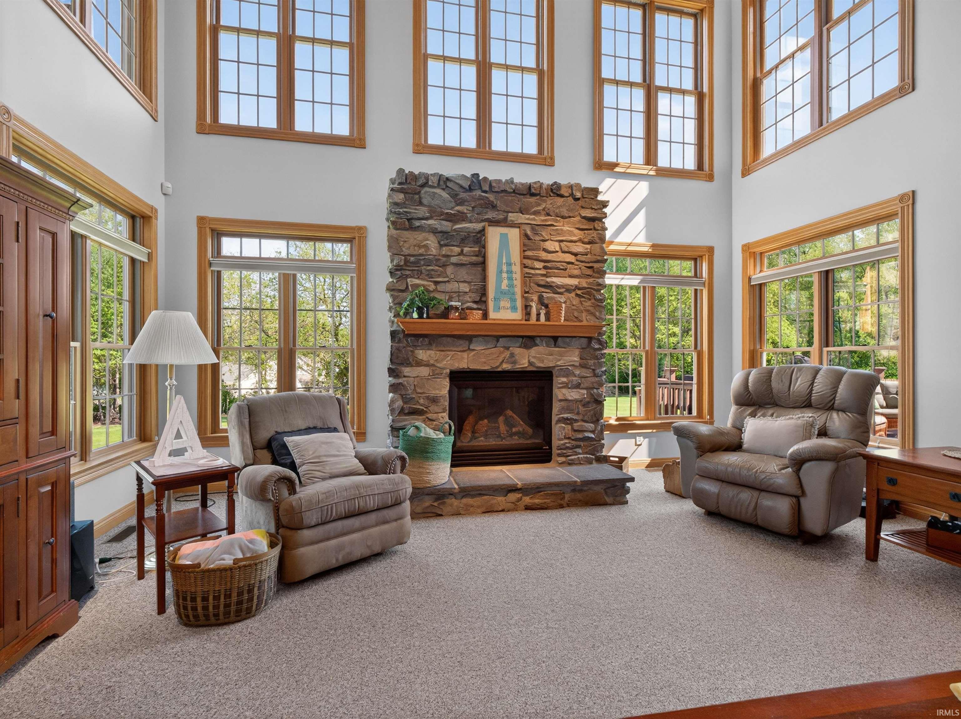 Two-story living area featuring a stone fireplace with a wood mantle, numerous windows with grid patterns, light-toned carpeting, and wood-finish trim