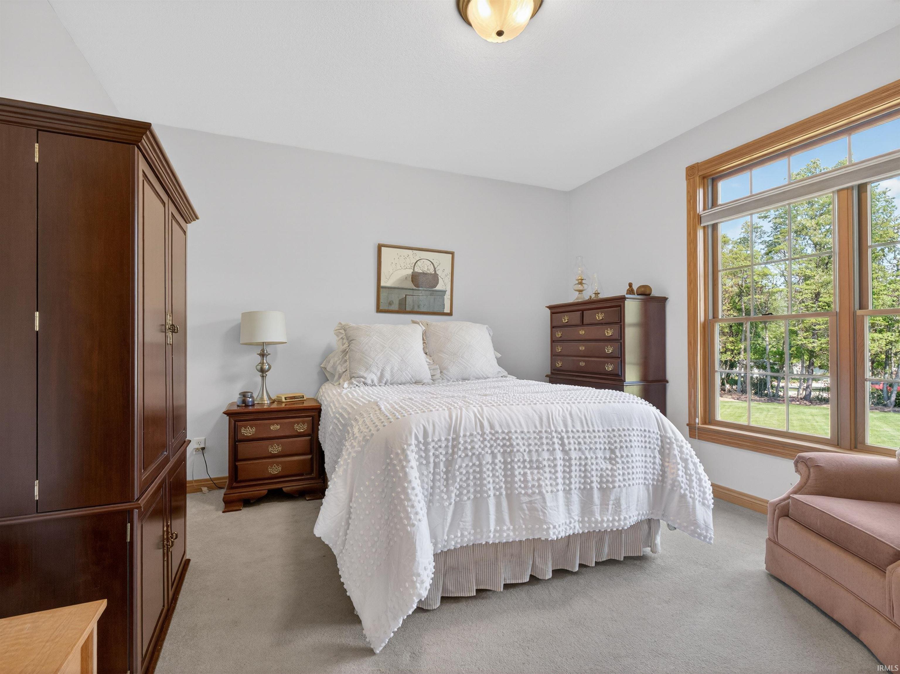 Bright bedroom featuring a large window with natural wood trim, plush carpet flooring, a neutral wall palette, and a semi-flush mount ceiling light fixture