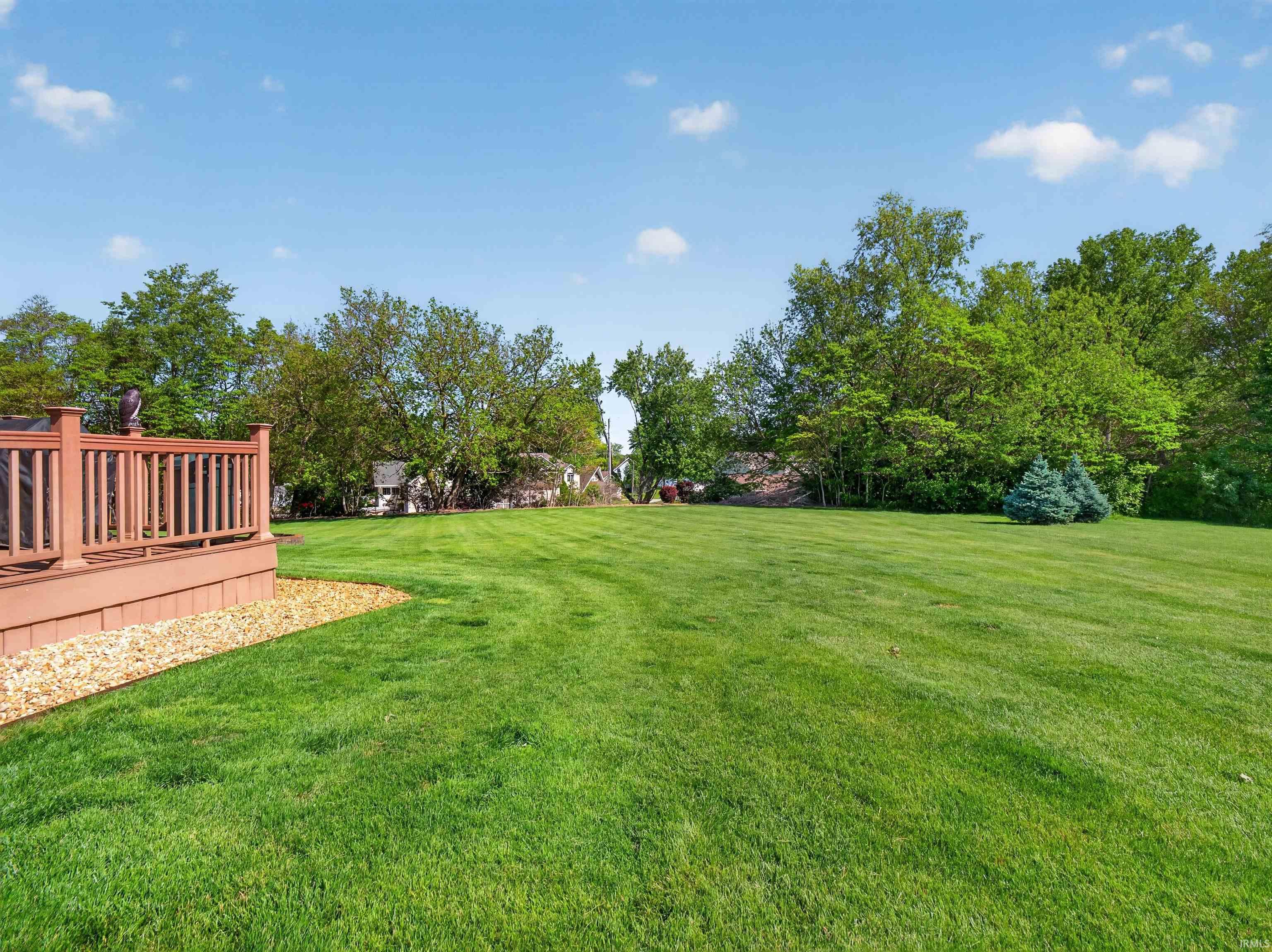 Expansive rear yard featuring a wooden deck with railing, a gravel border, and mature perimeter trees