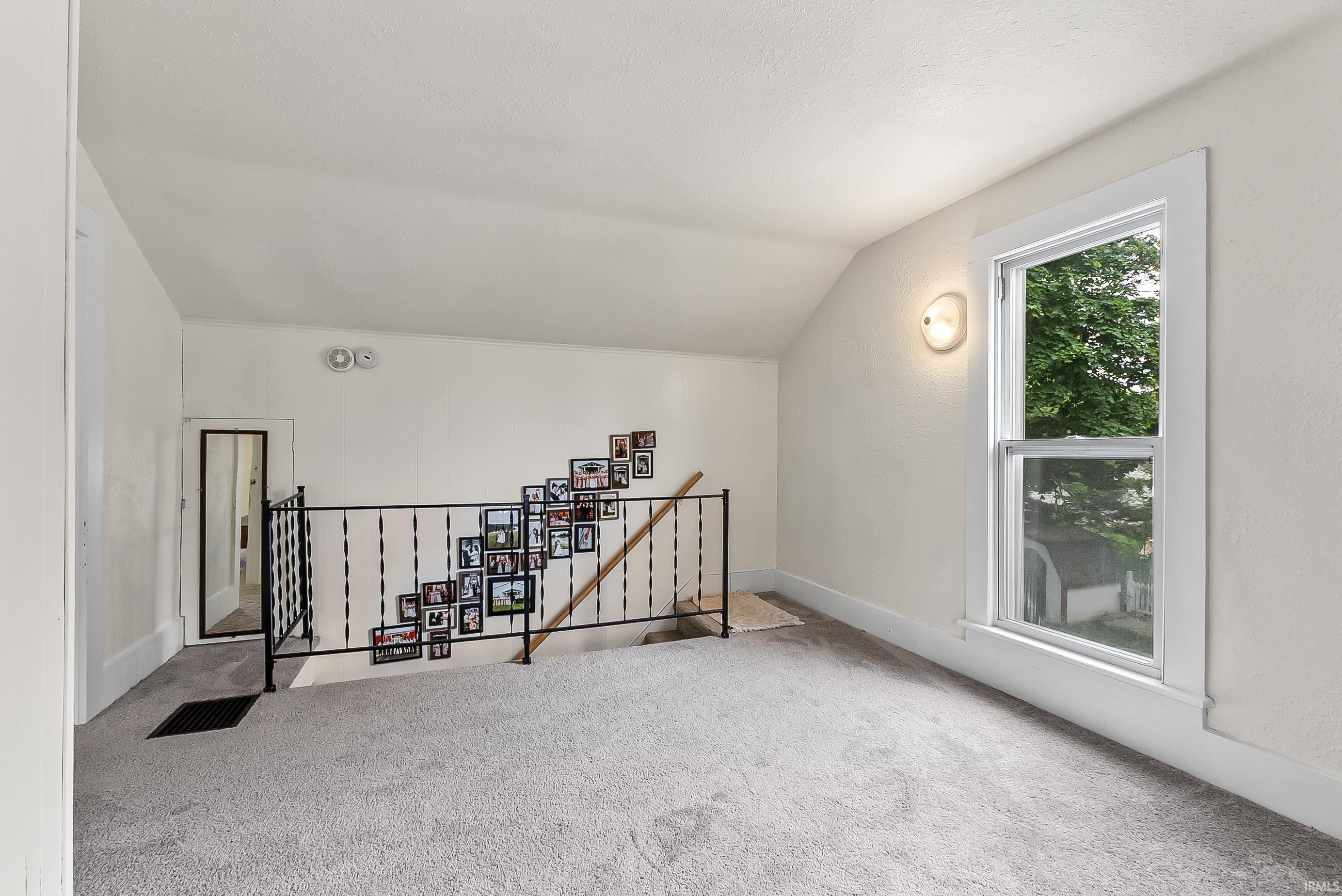 Carpeted upper-level space featuring a single-hung window with white trim, a built-in wall sconce, and a black wrought iron stair railing