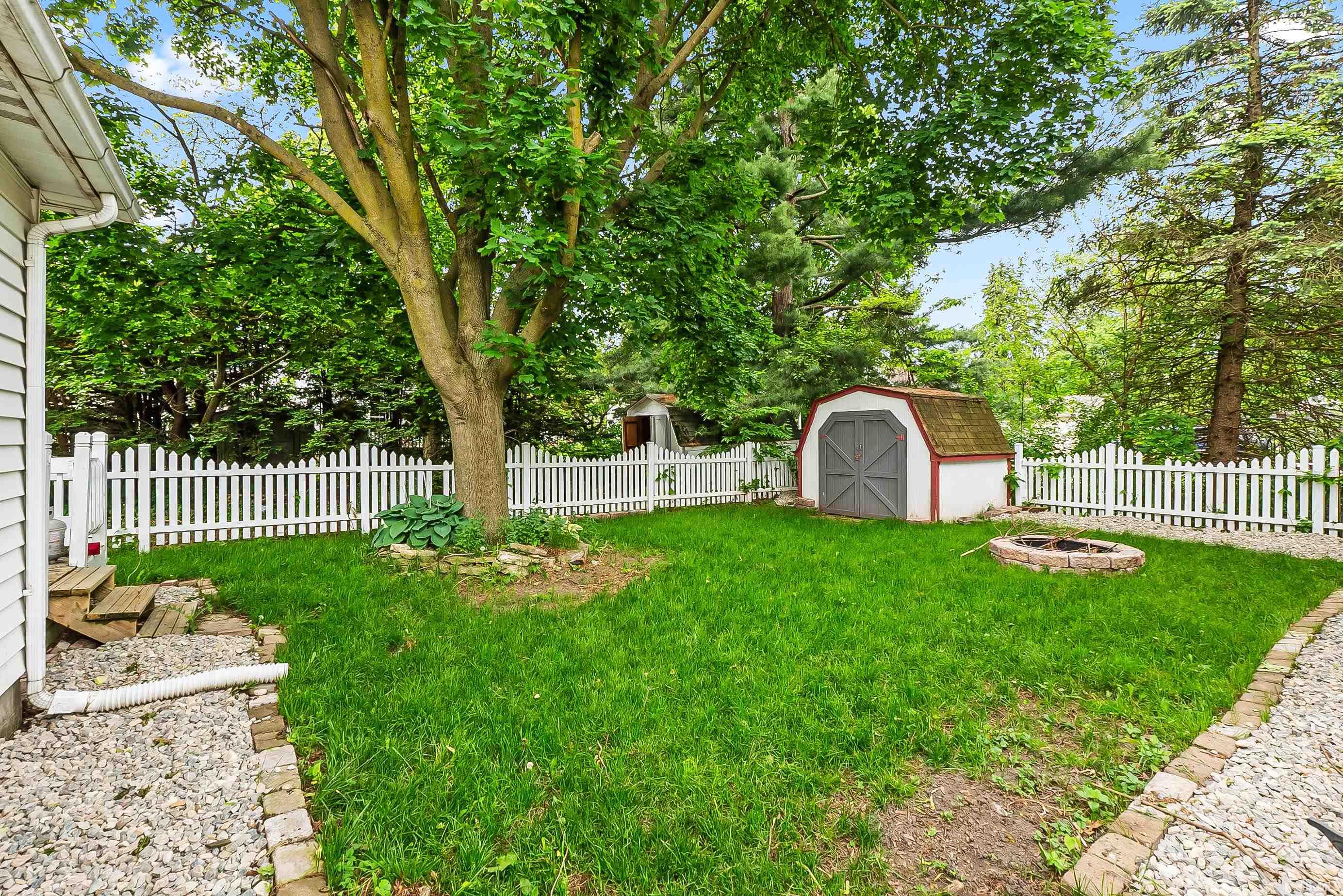 Fenced rear yard featuring a lawn, white picket fencing, mature trees, a garden shed, and a stone-lined fire pit