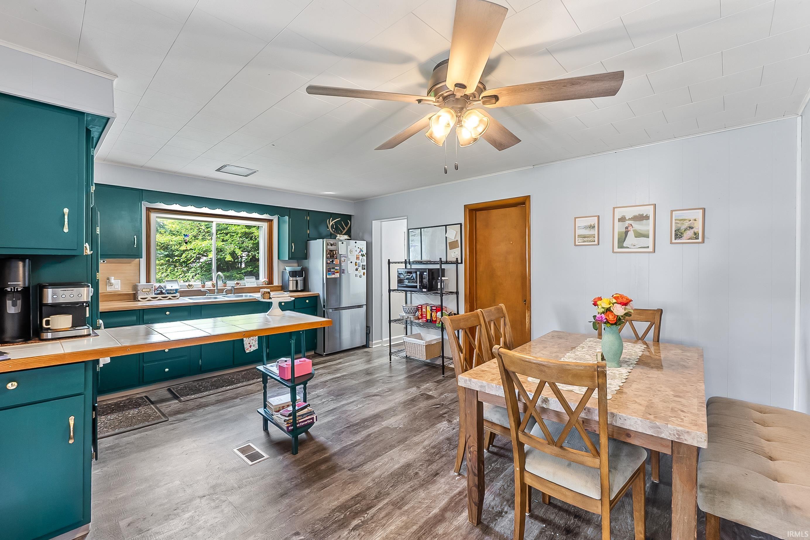 Kitchen and dining area featuring deep teal cabinetry, wood-finish flooring, a stainless steel refrigerator, a double-basin sink, and a ceiling fan with integrated lighting