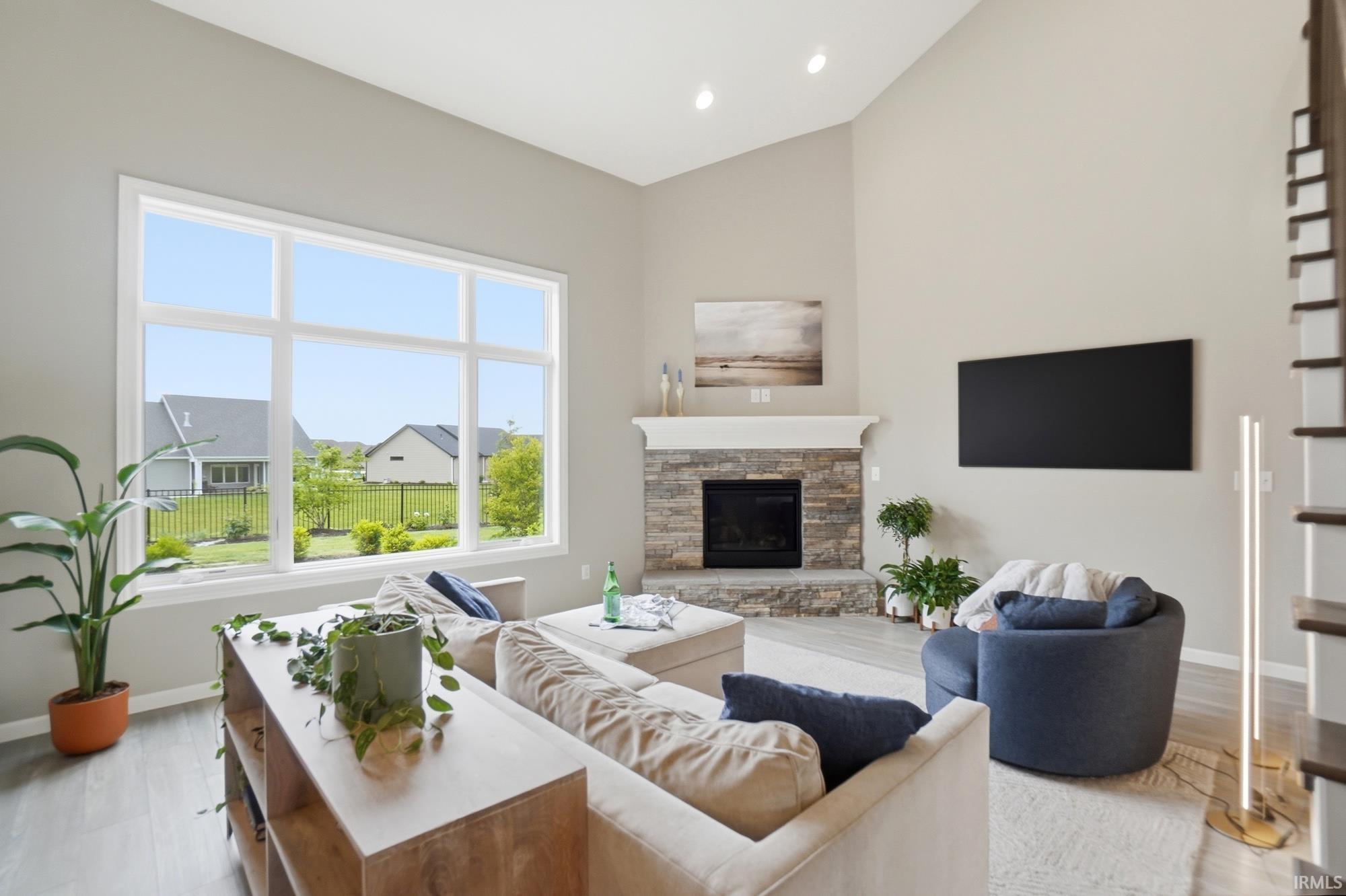 Living area featuring a stone-clad fireplace with a white mantel, large multi-pane windows, vaulted ceilings, and light wood-finish flooring