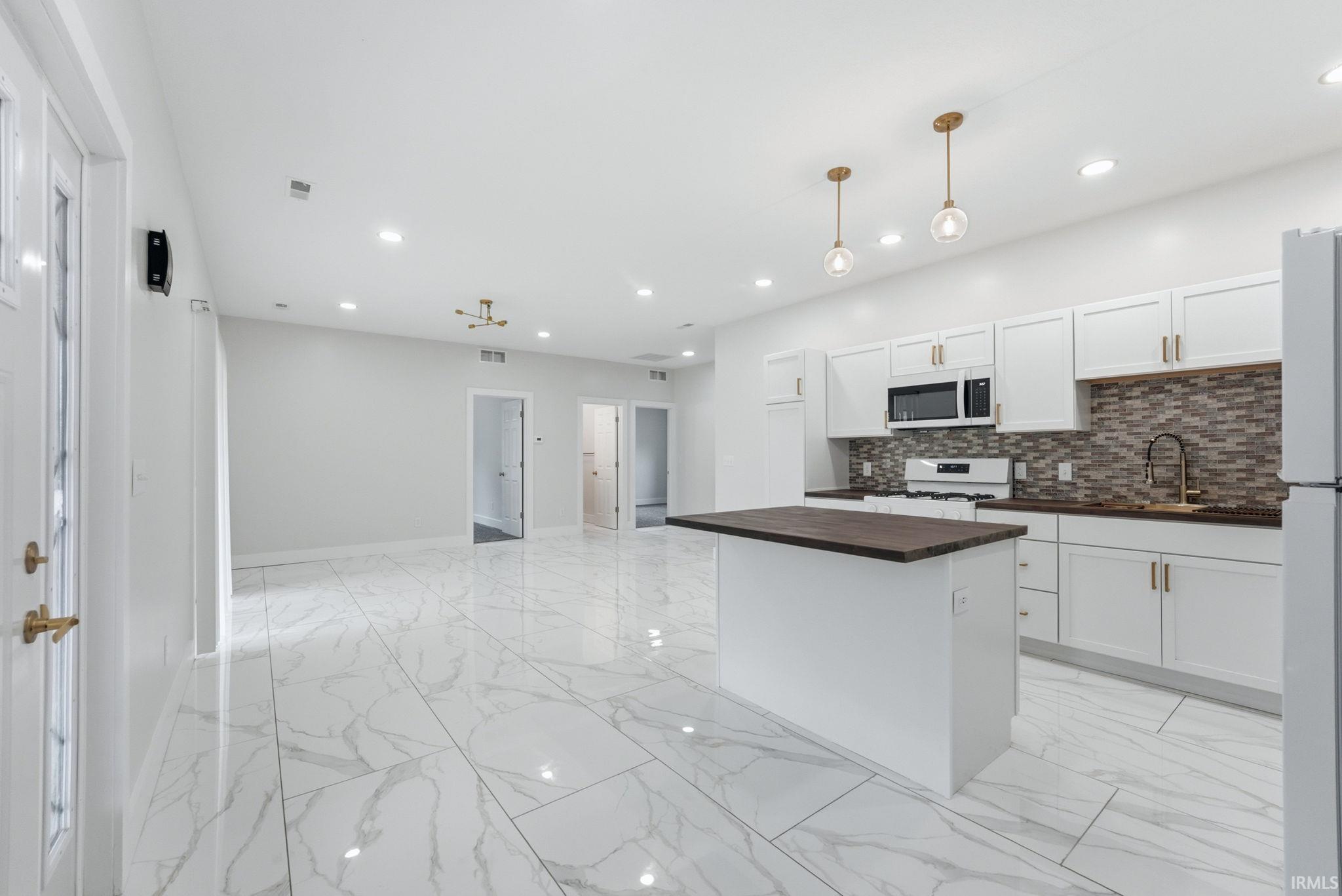 Open-concept kitchen and living area featuring marble-look tile flooring, white shaker cabinetry with gold-tone hardware, a dark wood-finish kitchen island, a mosaic tile backsplash, and recessed lighting
