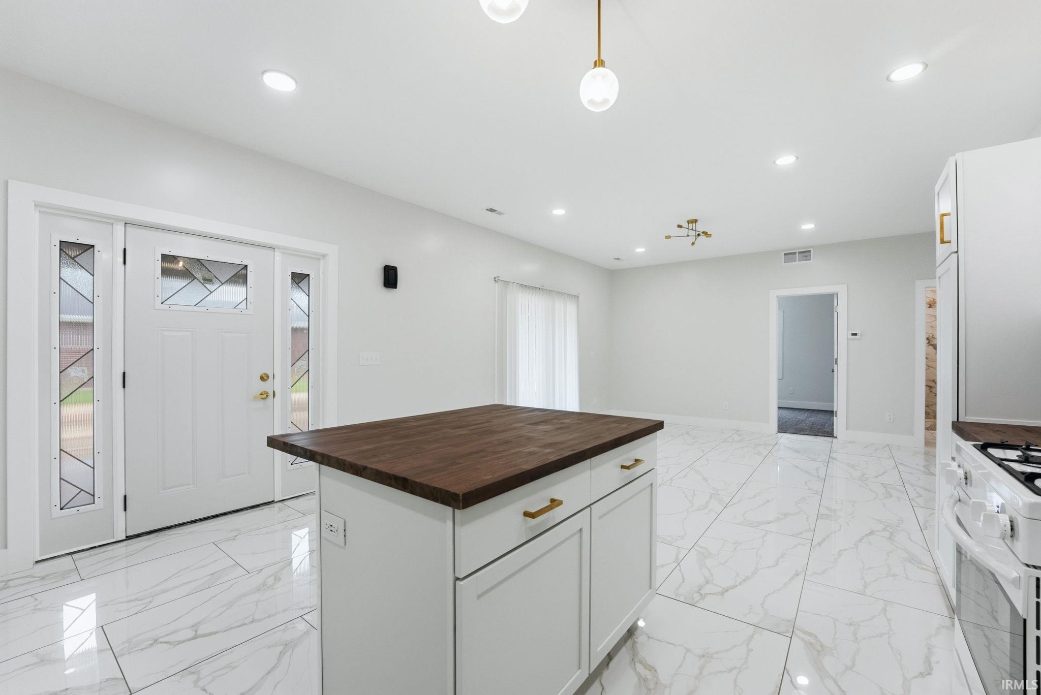 Entryway featuring a white door with decorative glass inserts and side panels, a kitchen island with a wood-finish countertop, white cabinetry with gold-tone hardware, white marble-look tile flooring, and recessed lighting