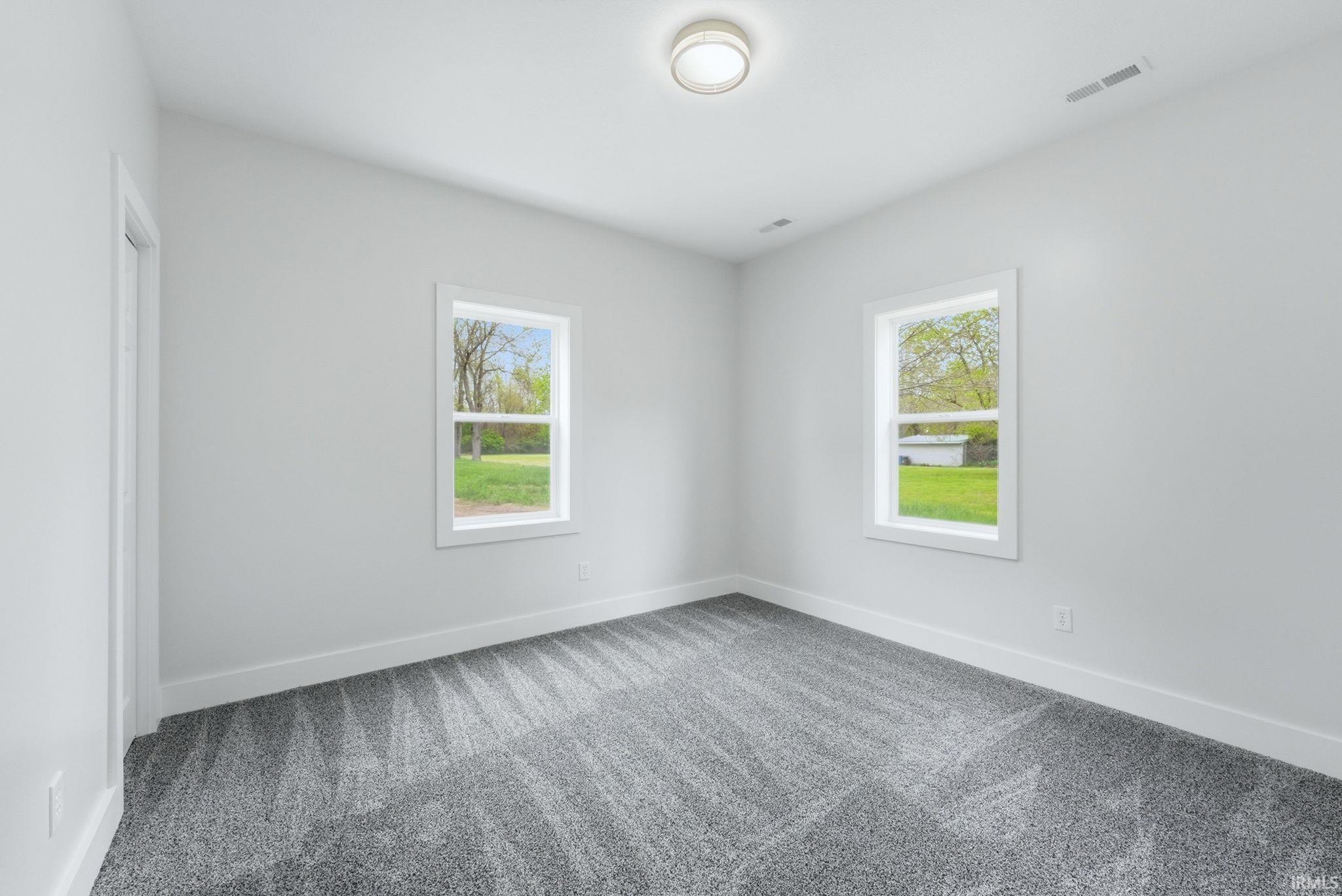 Bright room featuring two large windows, neutral gray wall tones, plush gray patterned carpeting, white trim, and a ceiling-mounted flush light fixture